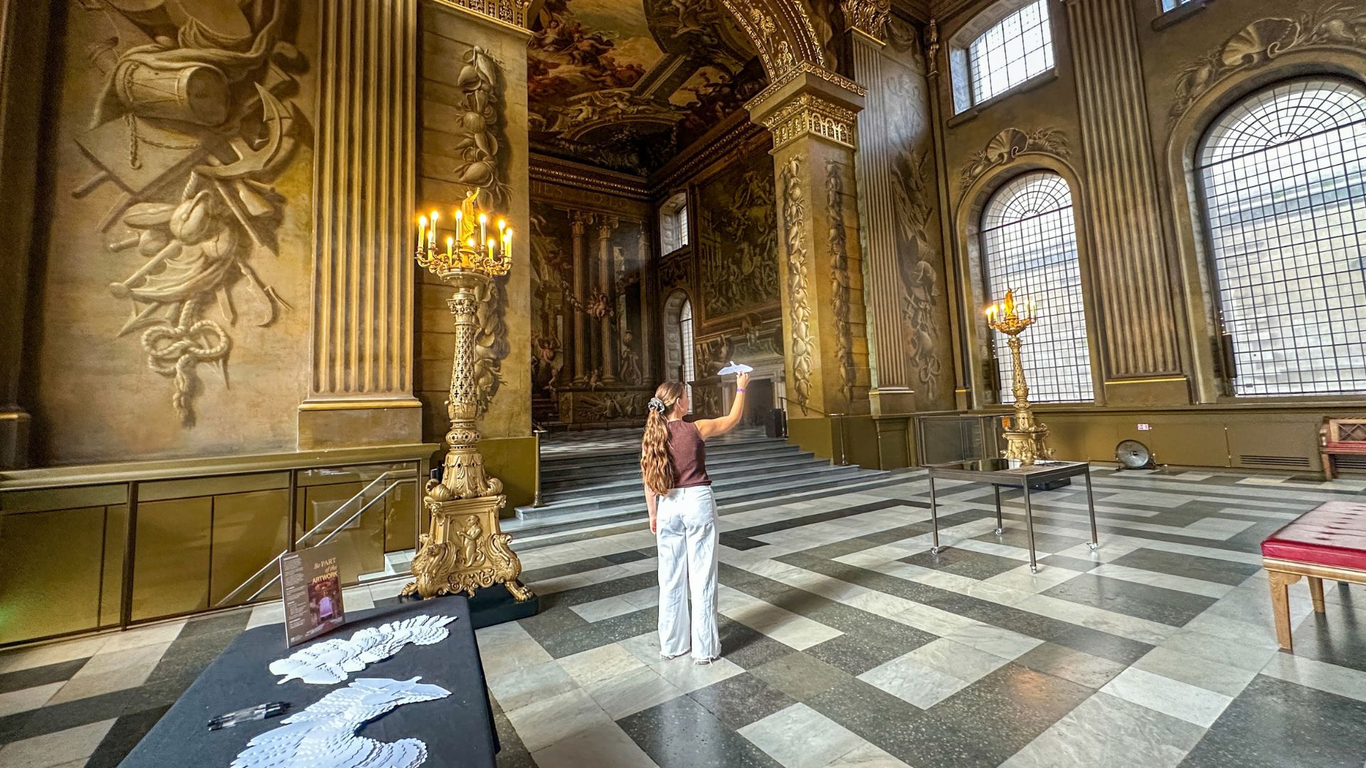 Explore the Painted Hall and get involved with the Peace Doves installation by creating your own paper dove. Image credit: Kirstine Spicer. A visitor stands inside the ornate Painted Hall beside a table displaying paper doves, surrounded by the hall's richly decorated walls and ceiling.