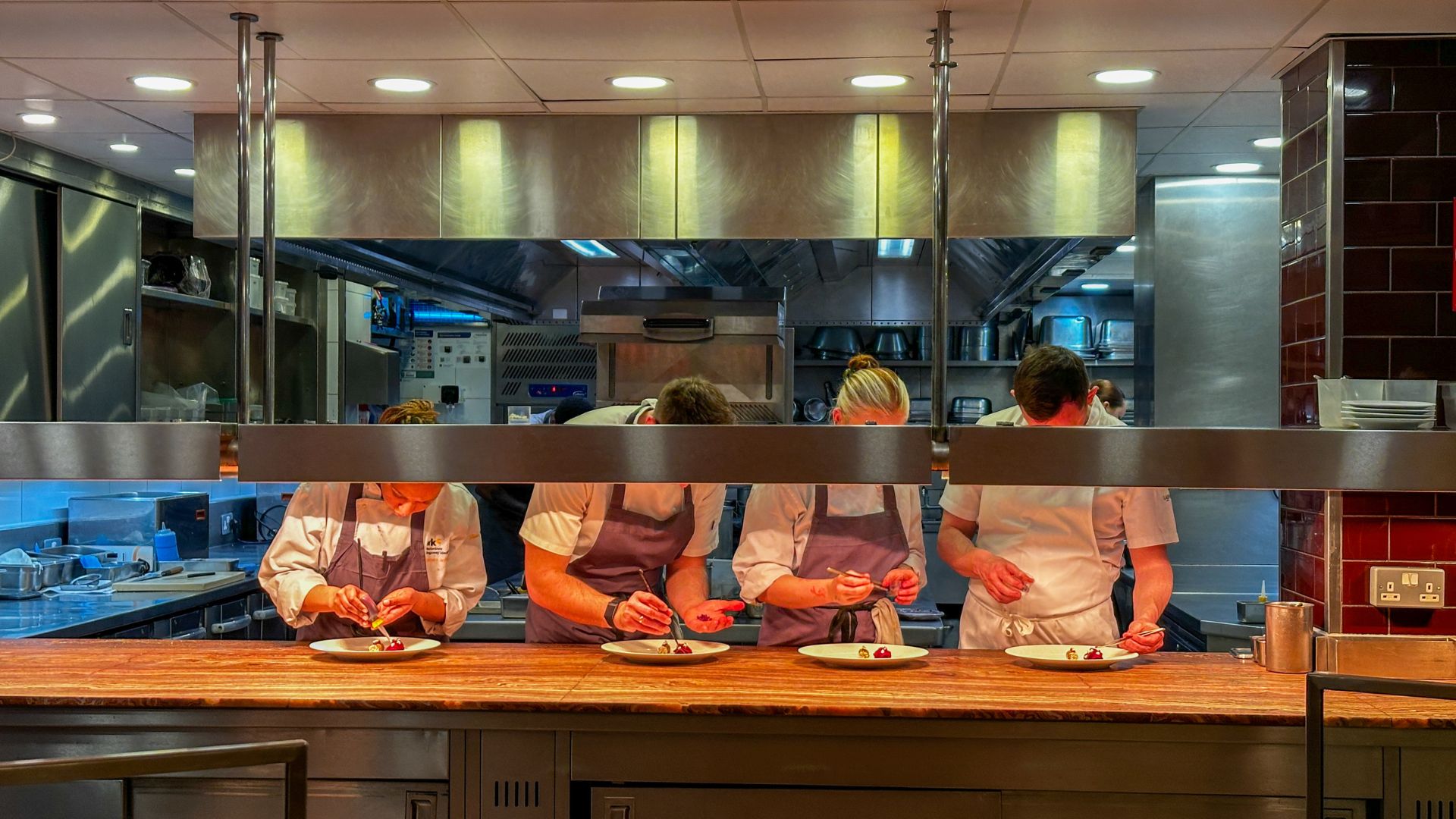 A row of chefs plating dishes simultaneously at the Pétrus kitchen pass beneath a stainless steel canopy.