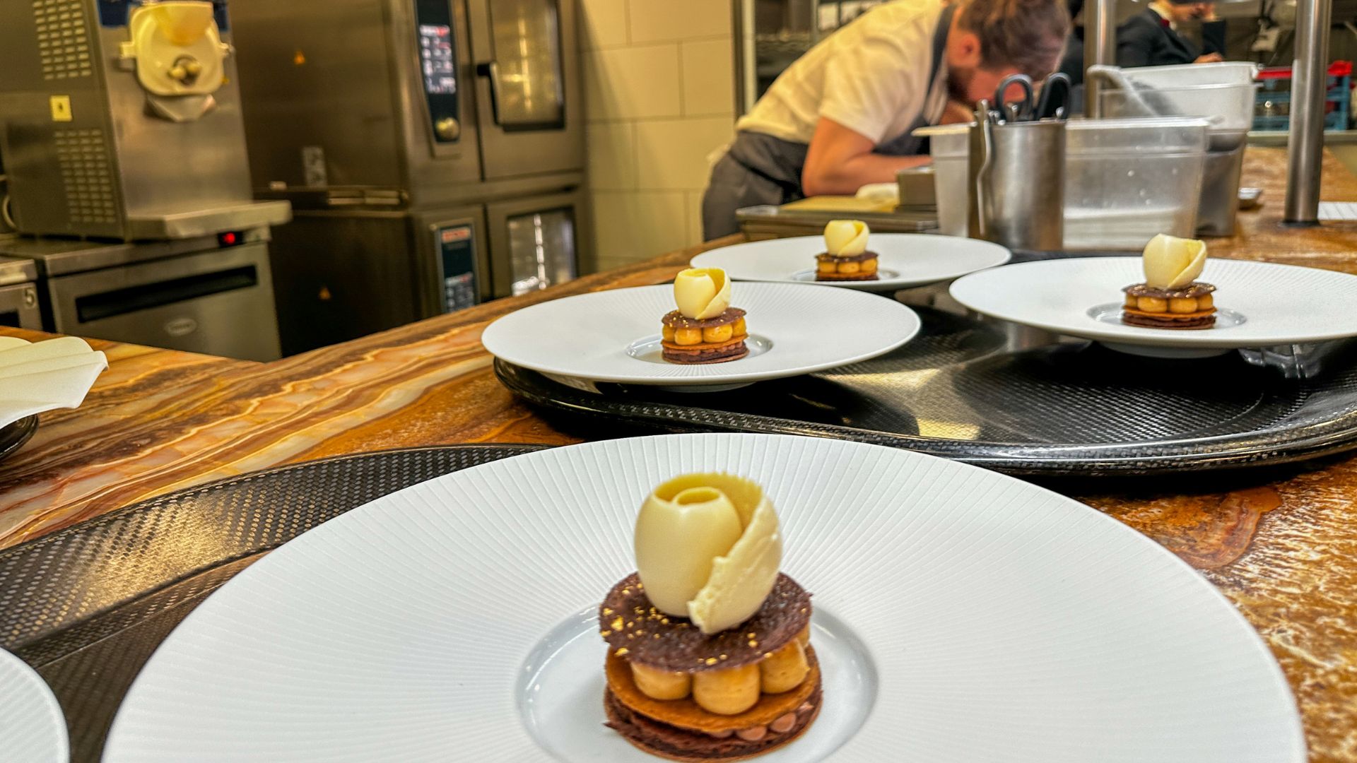 Multiple identical chocolate desserts topped with white chocolate roses lined up on the kitchen pass at Pétrus, with a chef working in the background.
