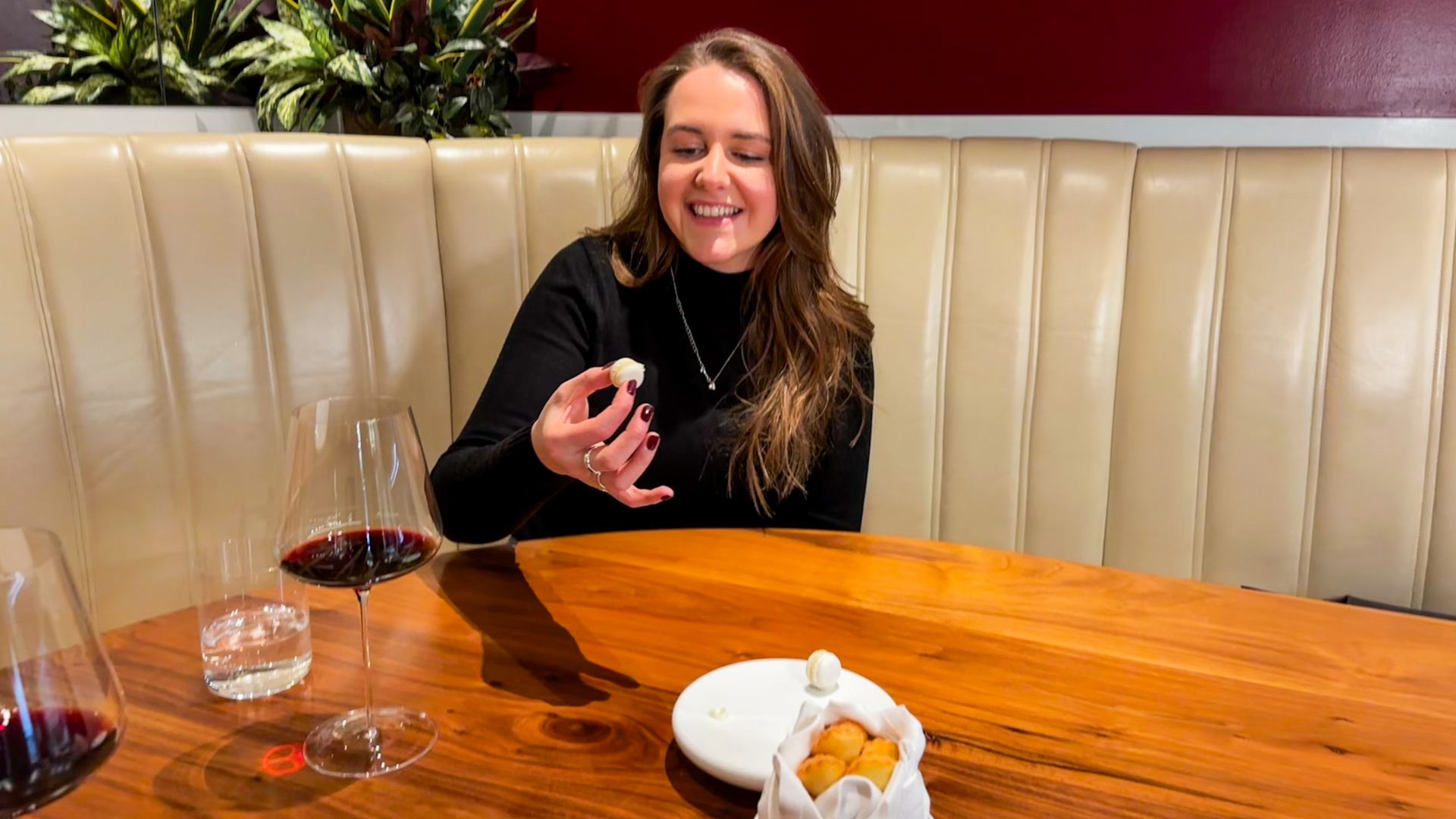 Diner seated at the Pétrus Kitchen Table smiling while holding a canapé, with wine glasses on the wooden counter.