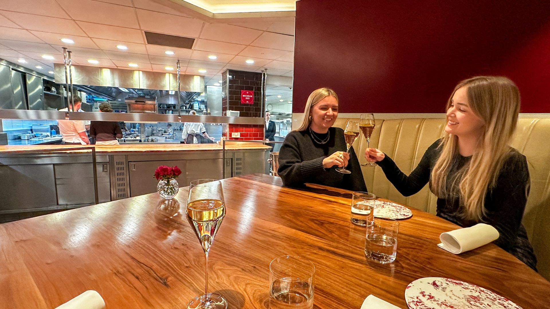 Two diners toasting glasses of sparkling wine at the Pétrus Kitchen Table with the open kitchen visible behind them.