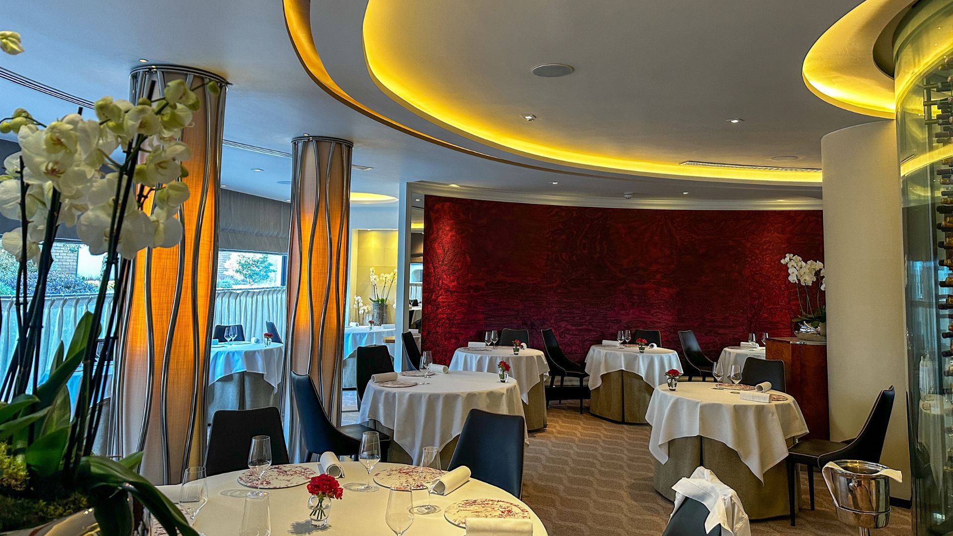 The softly lit dining room upstairs that feels calm, spacious and quietly luxurious. Photo credit: Visit London/Kirstine Spicer Elegant upstairs dining room at Pétrus with round white tablecloth tables, soft yellow ceiling lighting and a red feature wall.