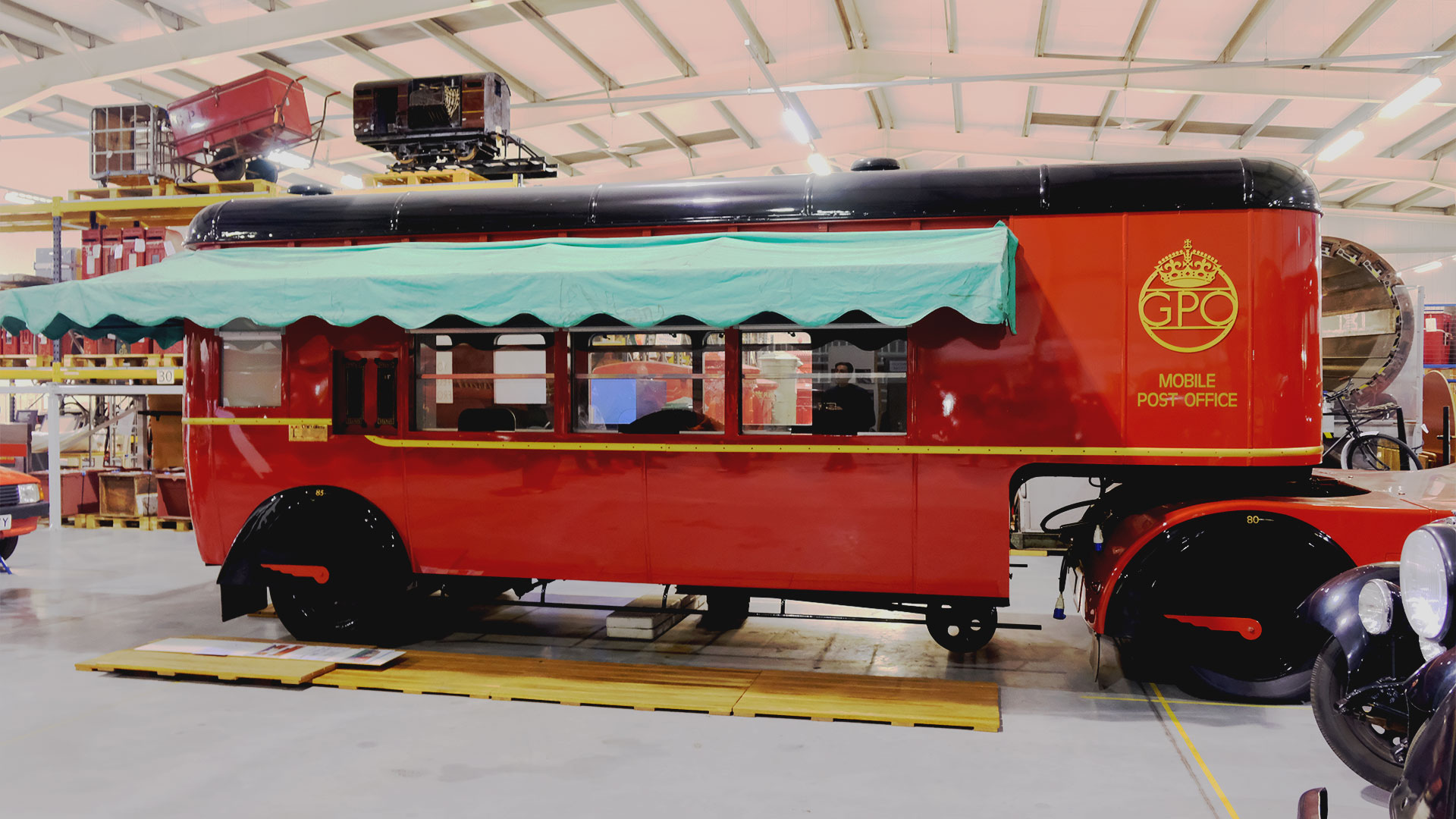 A large red mobile Post Office trailer with a green cover, acting as a post office complete with a phone booth and postal services, is on display at the Postal Museum Store. 