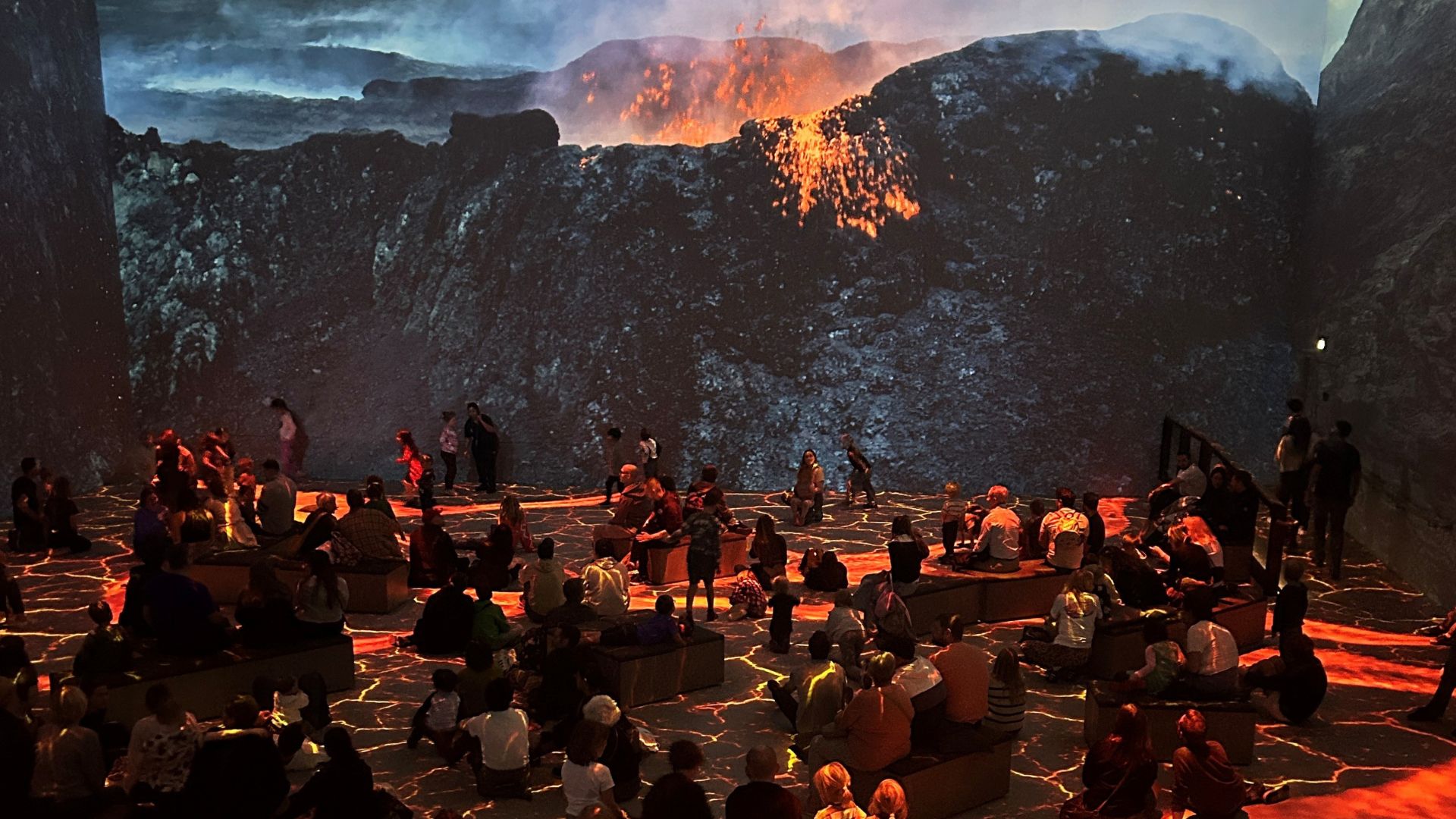 Watch an immersive volcanic eruption scene at Prehistoric Planet: Discovering Dinosaurs. Image courtesy of Hannah Cornish. People seated in a dark room with glowing lava patterns on the floor, facing a massive projection of an erupting volcano with smoke and molten rock.