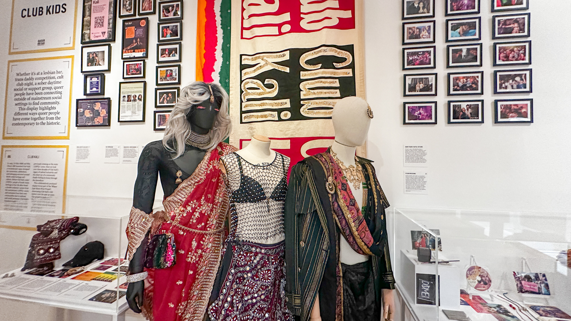 Three mannequins wearing bold, embellished club outfits stand in front of archival photographs and posters from London’s Club Kids scene.