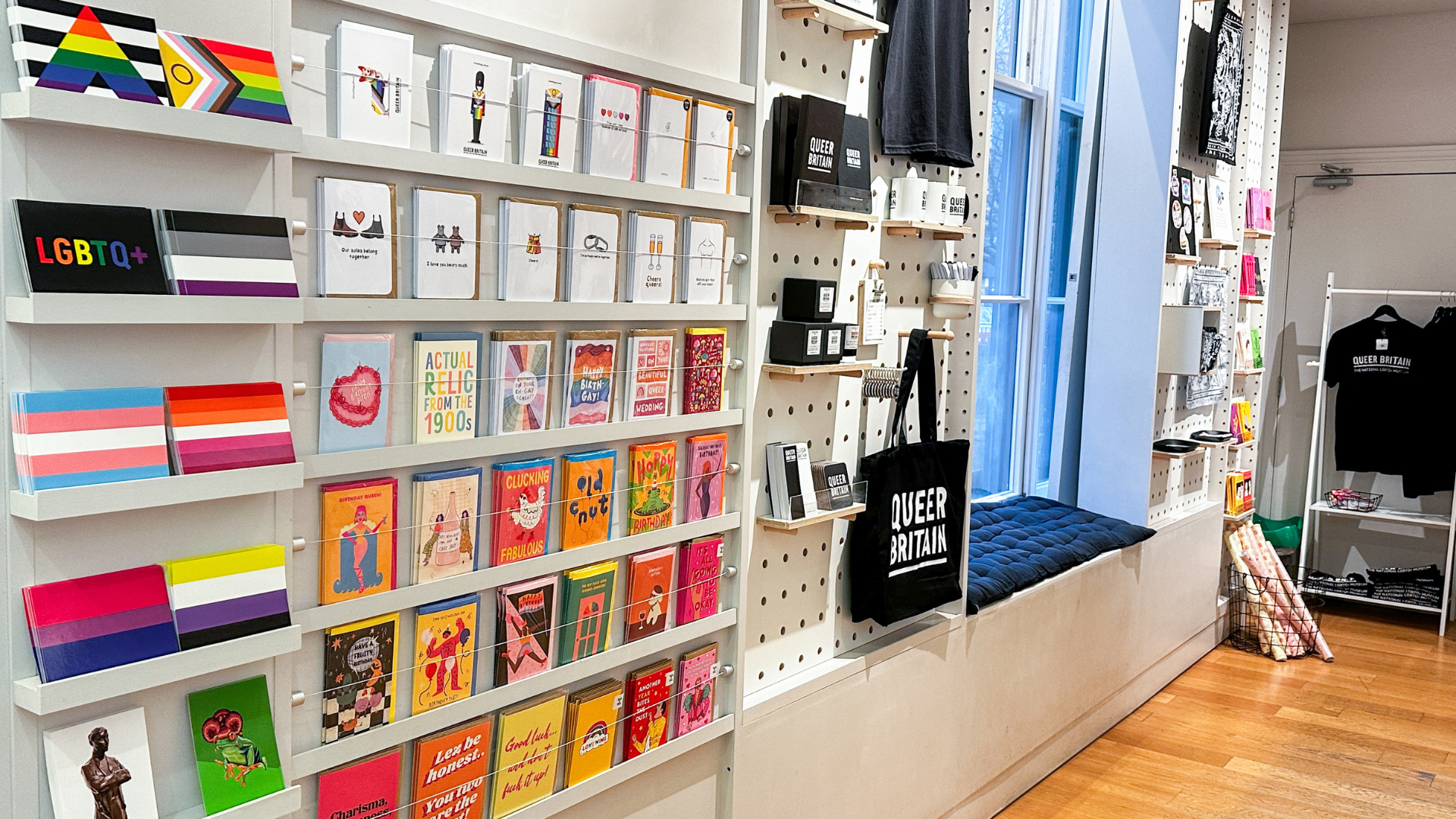 Shelves of LGBTQ+ themed books, cards and pride-flag stationery inside the Queer Britain Museum gift shop, with tote bags and apparel displayed nearby.