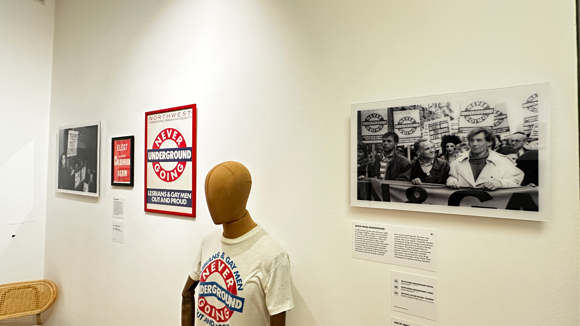 A mannequin wearing a “Never Going Underground” T-shirt stands beside framed protest posters and black-and-white photographs documenting LGBTQ+ activism.