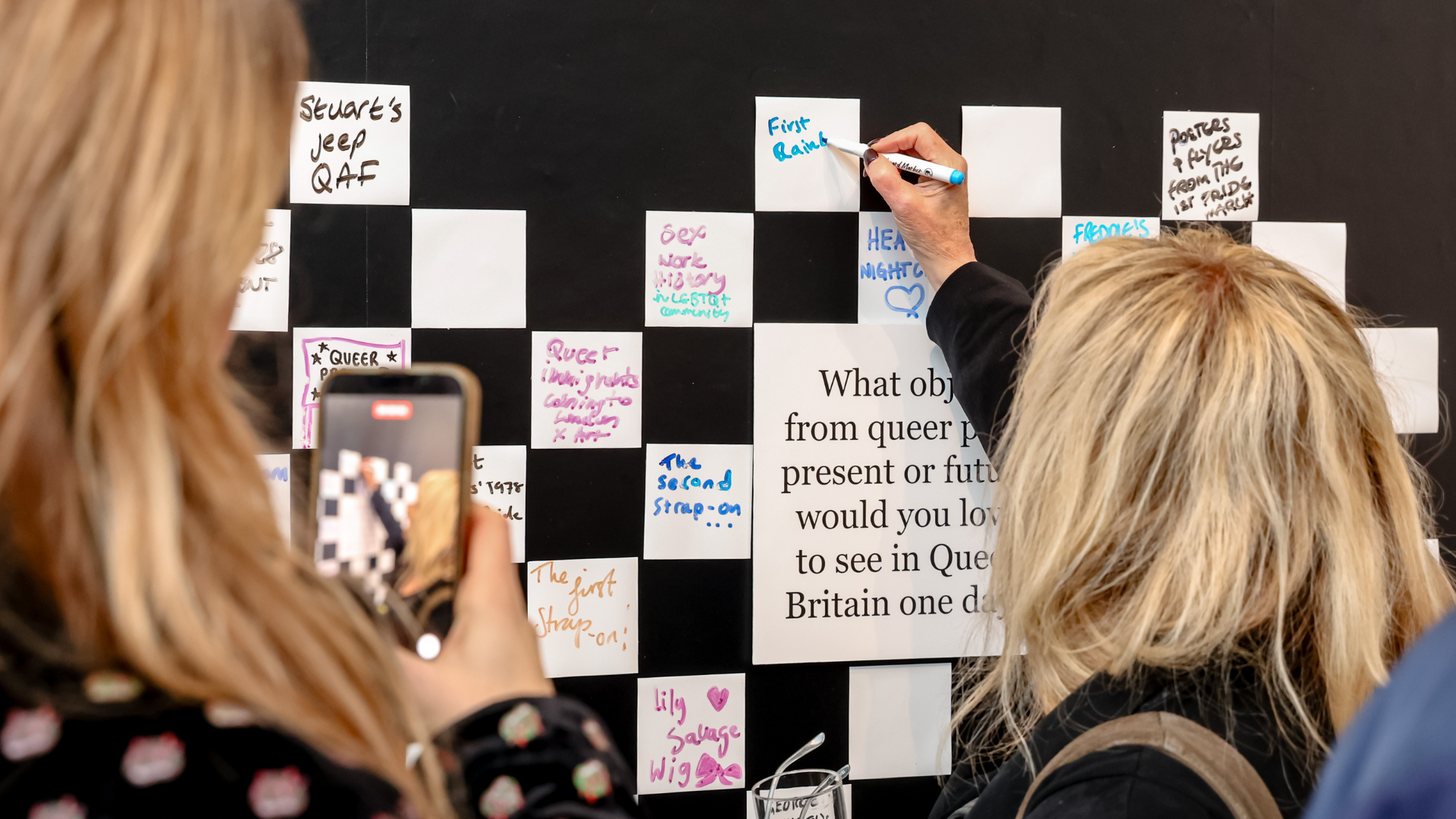 A wall of posters charting the visual history of BFI Flare (formerly London Lesbian & Gay Film Festival), showcasing decades of queer cinema, design and storytelling. Image courtesy of Queer Museum. Photo credit: Rahil Ahmad. Two visitors write notes on an interactive wall filled with handwritten messages responding to a question about queer history and objects.