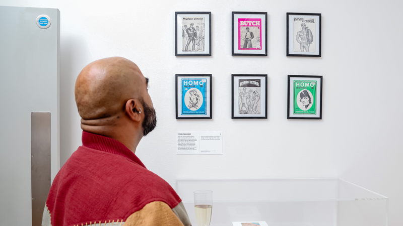 A museum visitor stands in front of framed magazine covers and illustrations displayed on a white gallery wall.