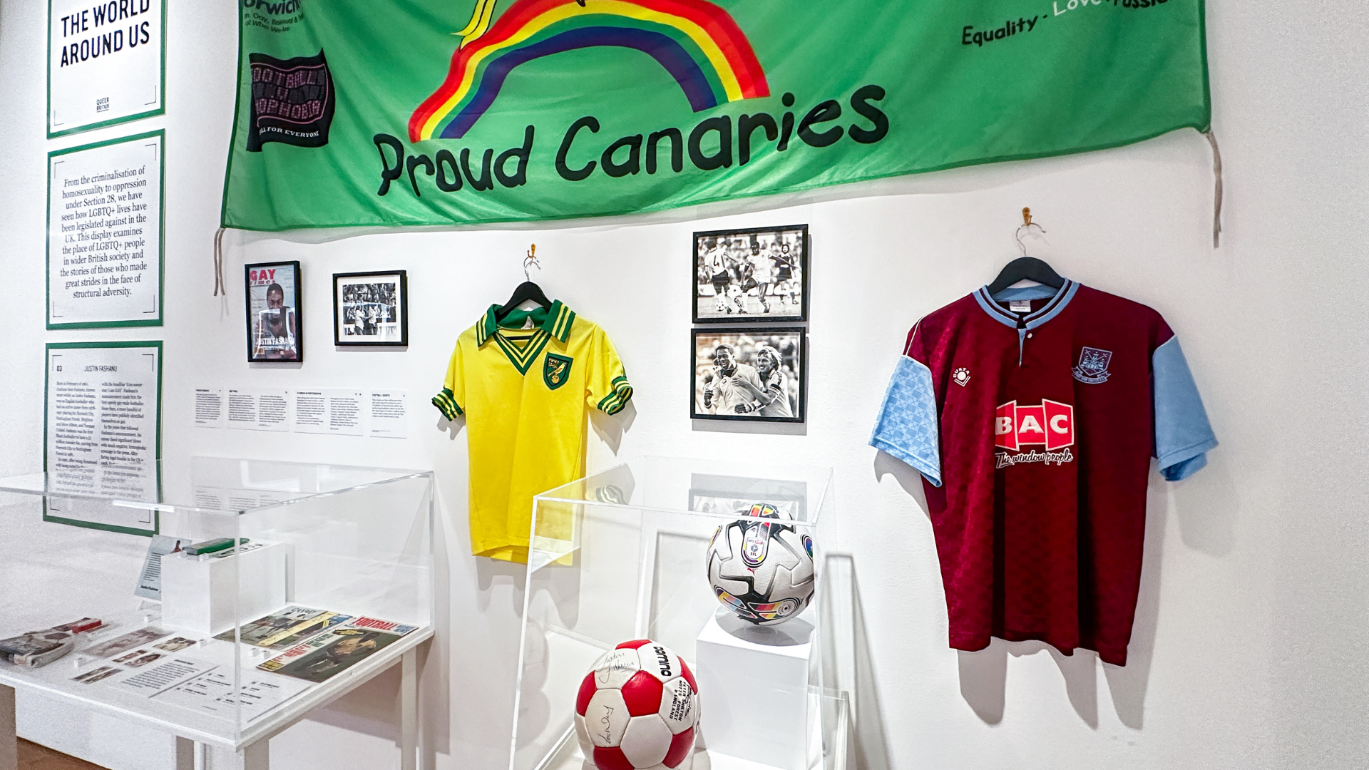 A museum display featuring football shirts, footballs and photographs beneath a green “Proud Canaries” banner with a rainbow, highlighting LGBTQ+ identity and sport.