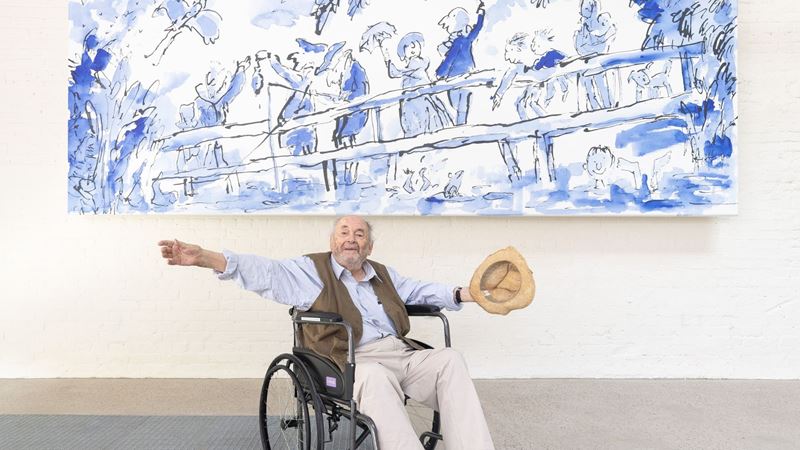 Quentin Blake sits in a wheelchair with his arms outstretched beneath a large blue-and-white illustration at the Quentin Blake Centre for Illustration. The artwork above depicts people gathered along a bridge, painted in Blake’s signature expressive ink and watercolour style.