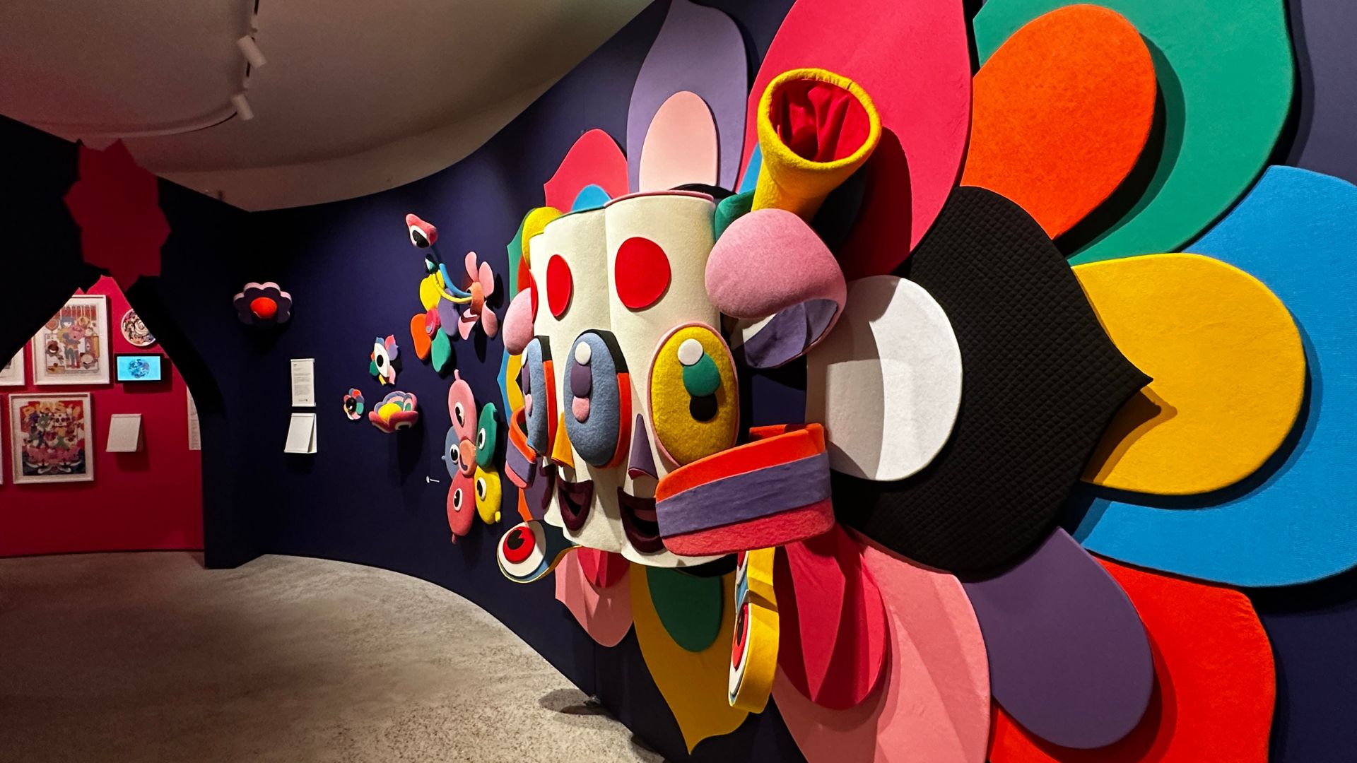 The striking MURU installation stops visitors in their tracks, transforming the gallery into an immersive world of colour and imagination. Photo Credit: Kirstine Spicer/Visit London. Large colourful MURU installation by MURUGIAH featuring layered shapes, flowers and mask-like forms.