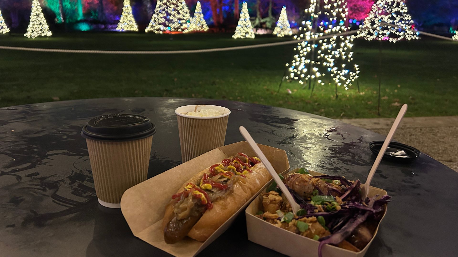 Table with hot drinks and festive food in front of illuminated Christmas trees in the garden