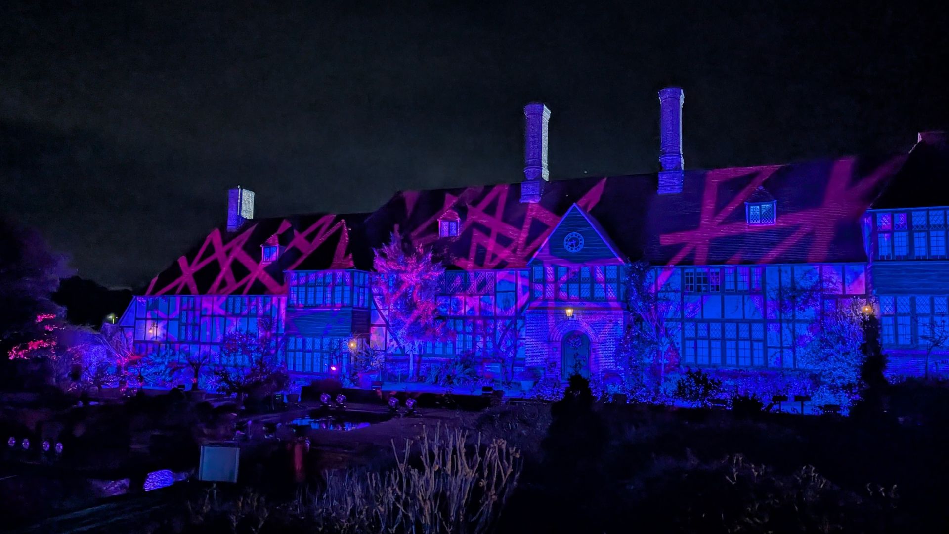RHS Wisley’s iconic building, Old Laboratory, transformed with dazzling light projections. Image courtesy of Hannah Cornish. Historic building facade lit with blue and pink crisscross light patterns