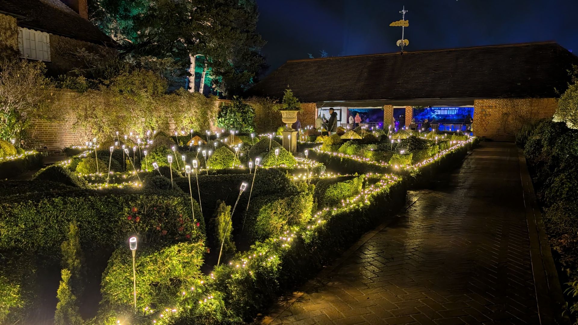 Elegant light displays highlight the beauty of the formal gardens at RHS Glow at Wisley. Image courtesy of Hannah Cornish. Formal garden with hedges and pathways outlined in warm white fairy lights