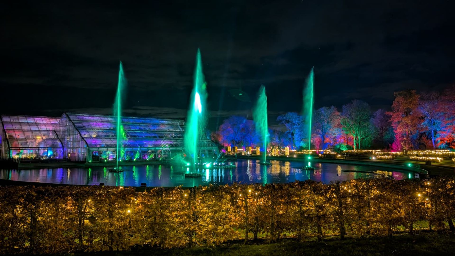 See the incredible floating pontoon at RHS Glow at Wisley, new for 2025. Image courtesy of Hannah Cornish. Night view of fountains lit in green with glasshouse and colourful trees in the background