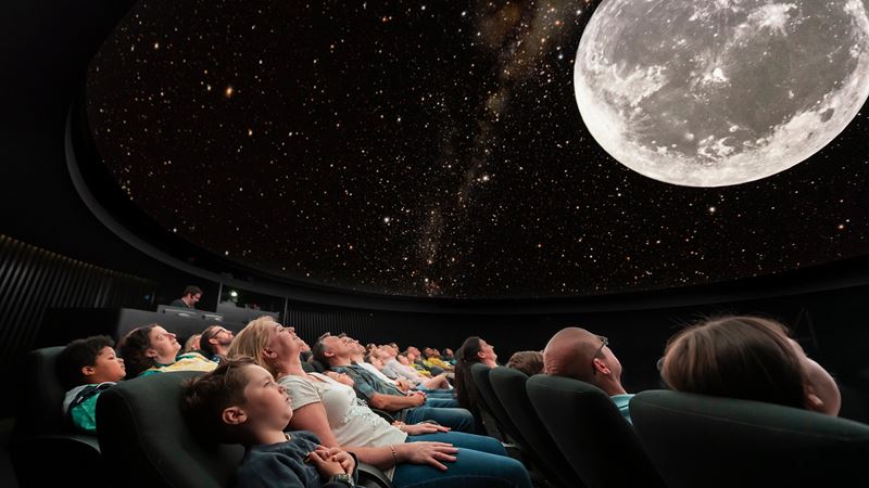 Audience members lay back in their seats, in the dark, looking up at a planetarium show which shows the moon and stars.