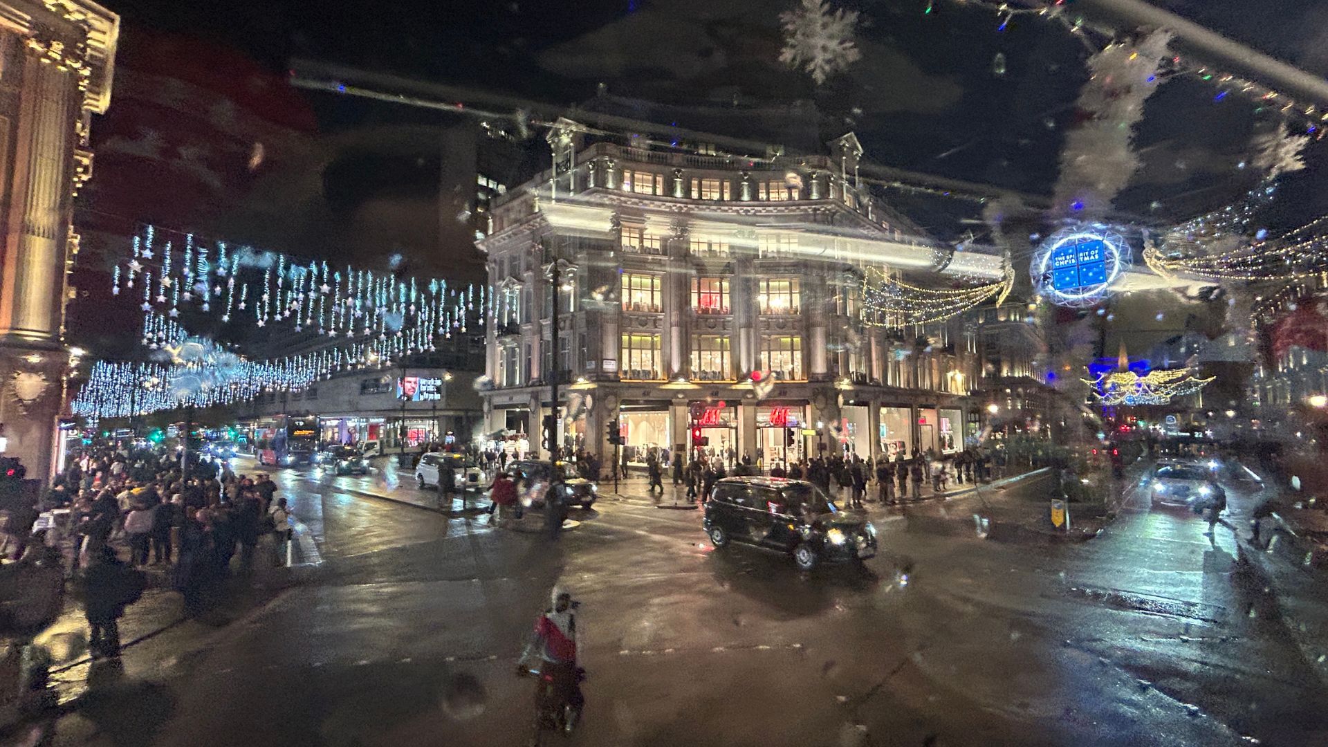 A busy London street at night with festive Christmas lights, including cascading blue lights and glowing shopfronts, viewed from a bus.
