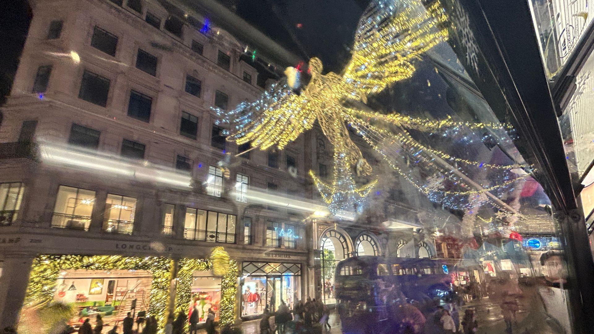 View through a bus window of Regent Street at night, showcasing illuminated angel-shaped Christmas lights and decorated shopfronts with glowing displays.