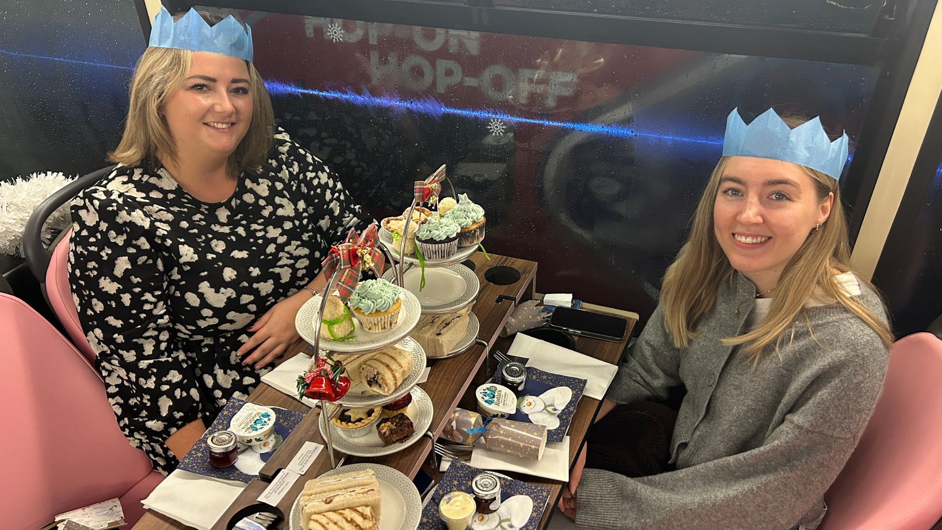 Pair a festive afternoon tea and London's iconic Christmas lights onboard the Snowman afternoon tea bus tour. Image courtesy of Hannah Cornish. A festive afternoon tea setup on a bus table featuring tiered stands with sandwiches, scones, cupcakes, and Christmas-themed decorations, with two people sat at the table wearing paper crowns.