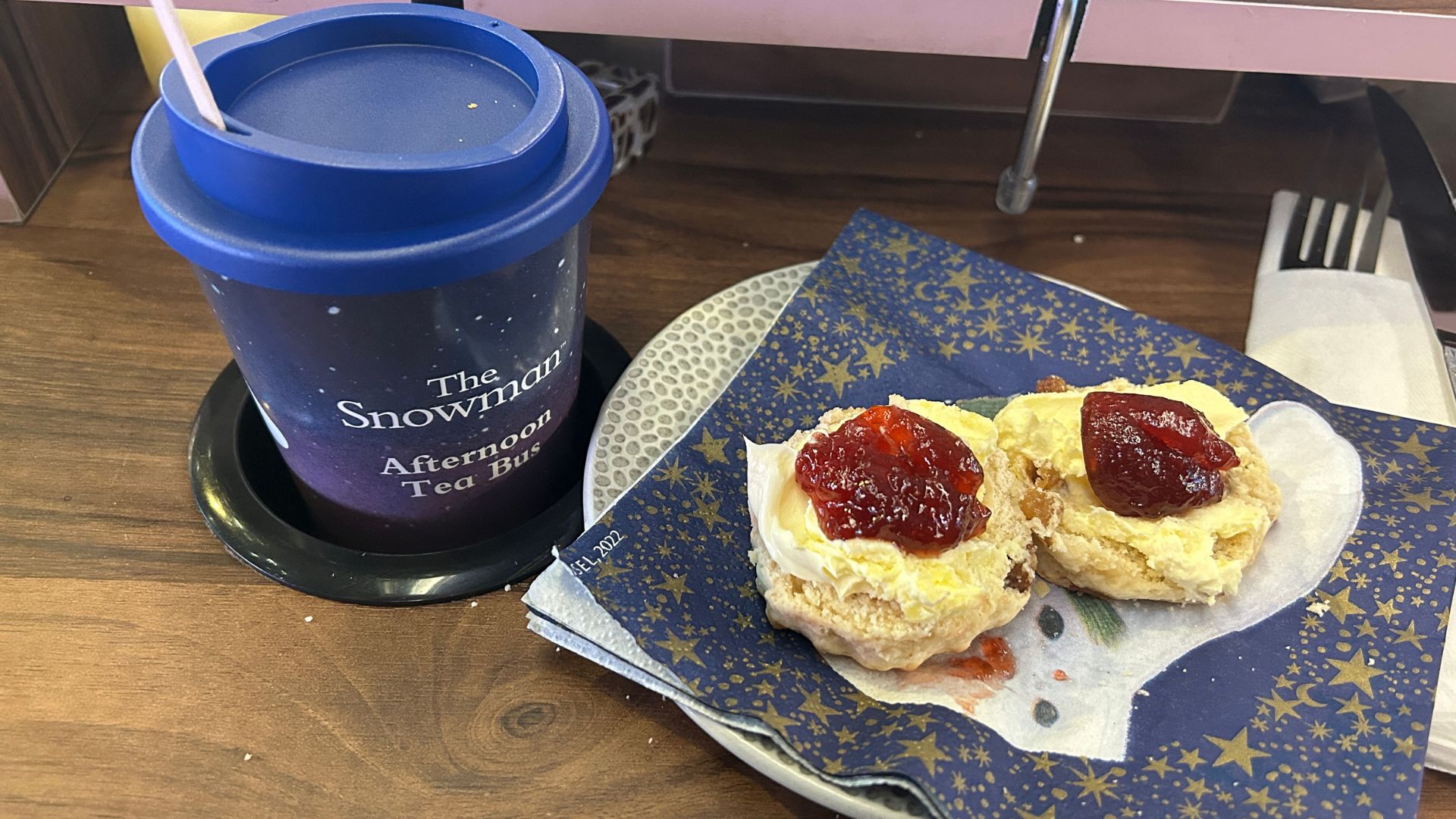 Enjoy warm scones with clotted cream and jam, and unlimited hot drinks served in a souvenir cup to take home at the end of the experience. Image courtesy of Hannah Cornish. Two scones topped with clotted cream and strawberry jam on a star-patterned napkin, next to a blue souvenir cup labelled “The Snowman Afternoon Tea Bus”