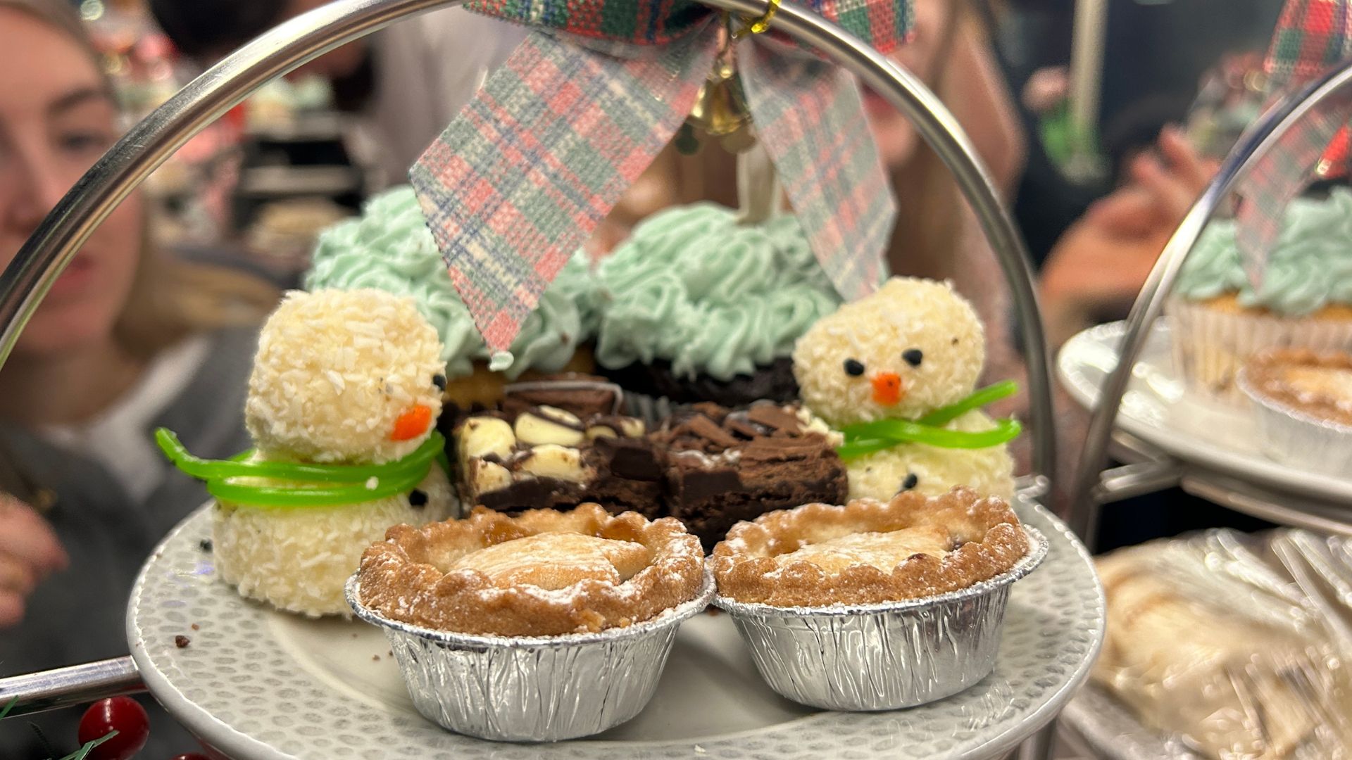Tuck into tasty festive treats including mini mince pies, gooey chocolate brownies and fluffy cupcakes onboard the Snowman afternoon tea bus tour. Image courtesy of Hannah Cornish. A close-up of a dessert tier featuring coconut snowman-shaped treats, chocolate brownies, and mince pies dusted with powdered sugar.