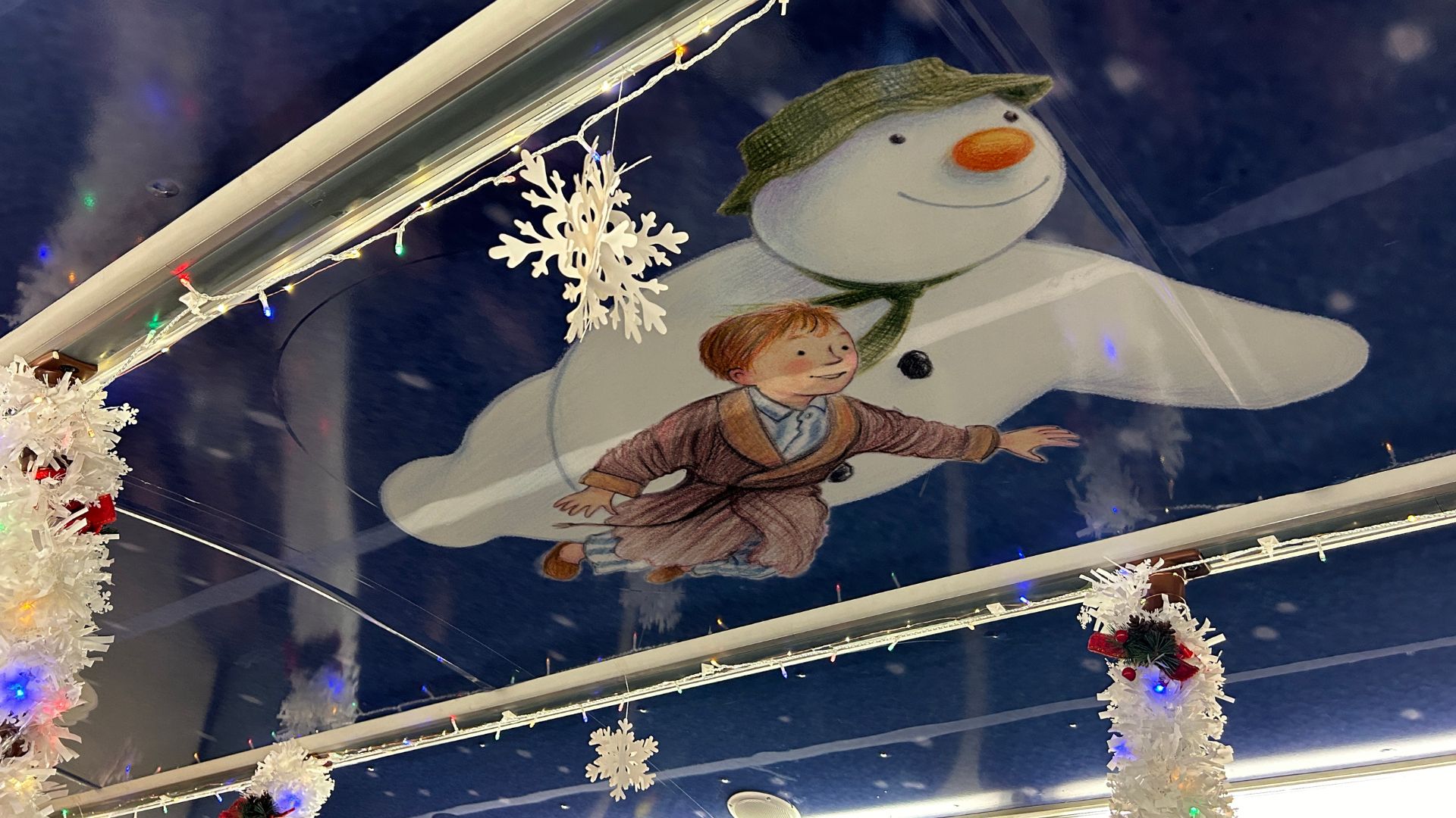 Ceiling decoration inside the bus featuring an illustration of The Snowman and a boy flying, surrounded by white garlands and snowflake ornaments.