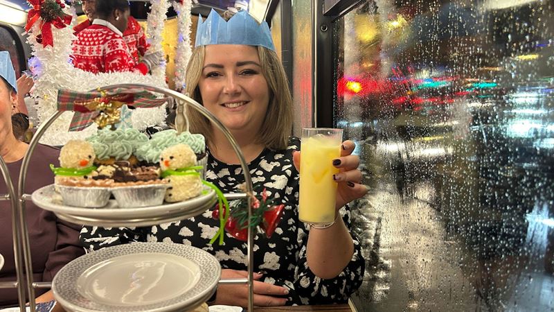 A festive bus table with a tiered stand of Christmas-themed treats, including snowman-shaped truffles and cupcakes, next to a person holding a glass of yellow drink.