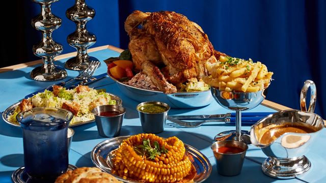 close up of a pale blue clothed dining table, with plates of rotisserie whole chicken, fries, dips and glasses of drink.