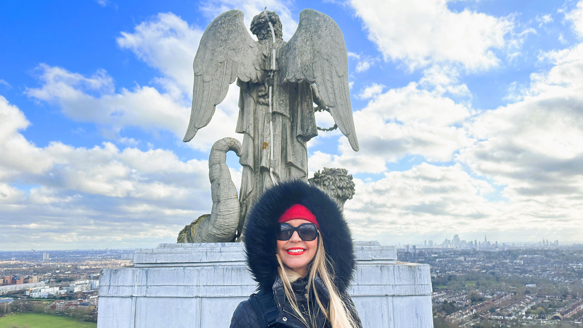 A girl with blonde hair wearing sunglasses and coat in front of the Angel of Plenty statue, admiring the views of London.