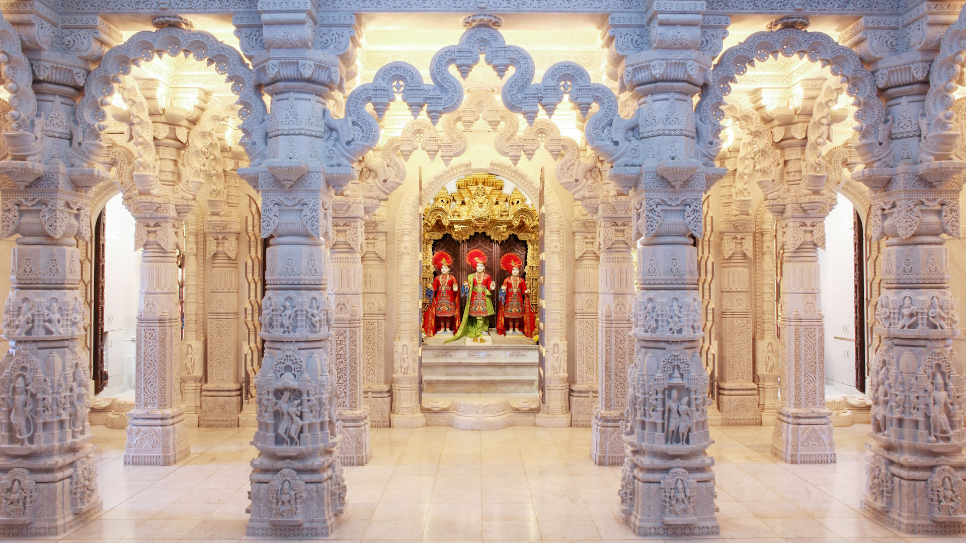 The garbh-gruh inner sanctum at BAPS Shri Swaminarayan Mandir. Image courtesy of BAPS Shri Swaminarayan Mandir. The garbh-gruh inner sanctum at BAPS Shri Swaminarayan Mandir with white limestone columns carved with deities and statues of deities.