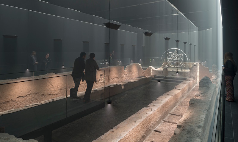 London Mithraeum uses light sculpture, haze and sound to bring the Temple of Mithras to life.. Photo credit: James Newton. Image courtesy of London Mithraeum Bloomberg Space. A couple walk through the remains of the Temple of Mithras, at the London Mithraeum.