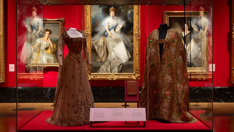Two dresses in a glass cabinet in the centre of an exhibition room with portraits of royals surrounding.