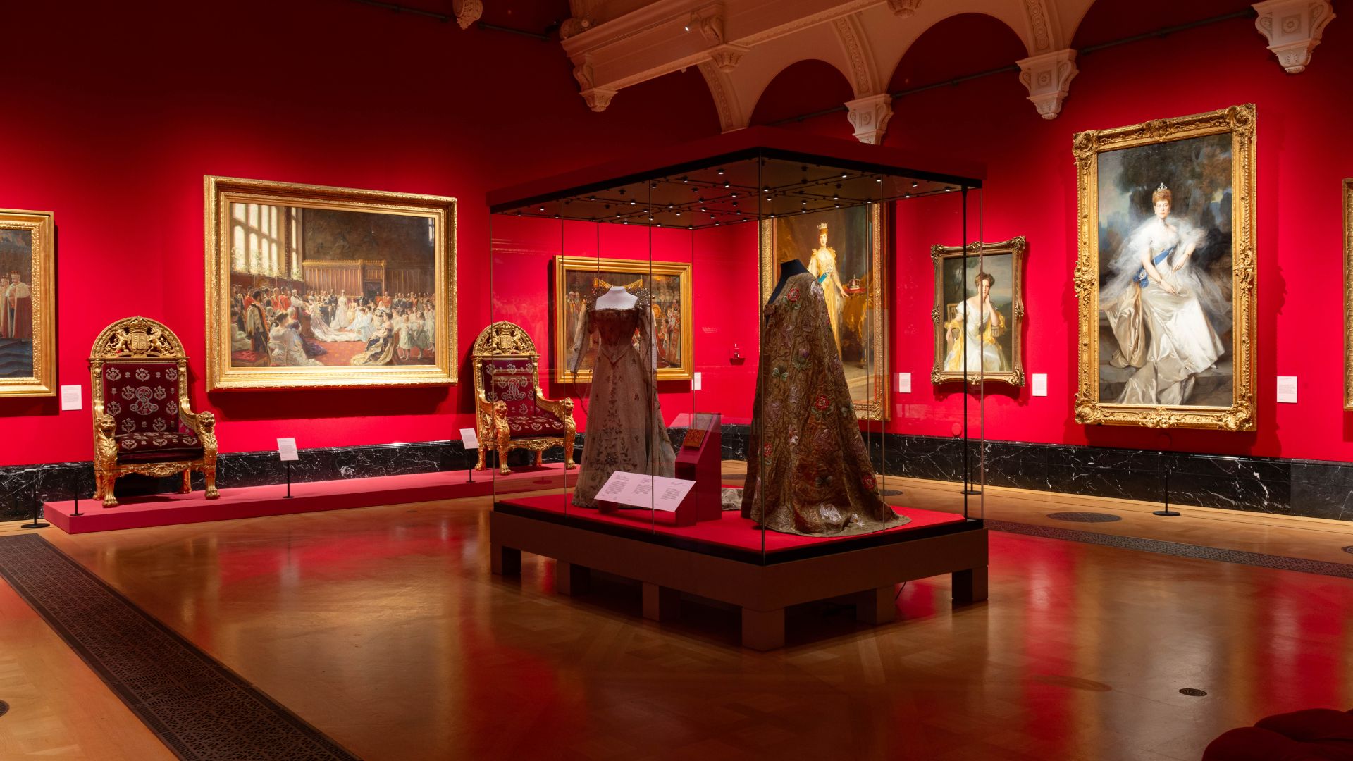 Delve into the glittering social lives of two sets of Kings and Queens at The King’s Gallery. © Royal Collection Enterprises Limited 2025 | Royal Collection Trust. Coronation dresses in centre of red gallery room with several paintings surrounding.