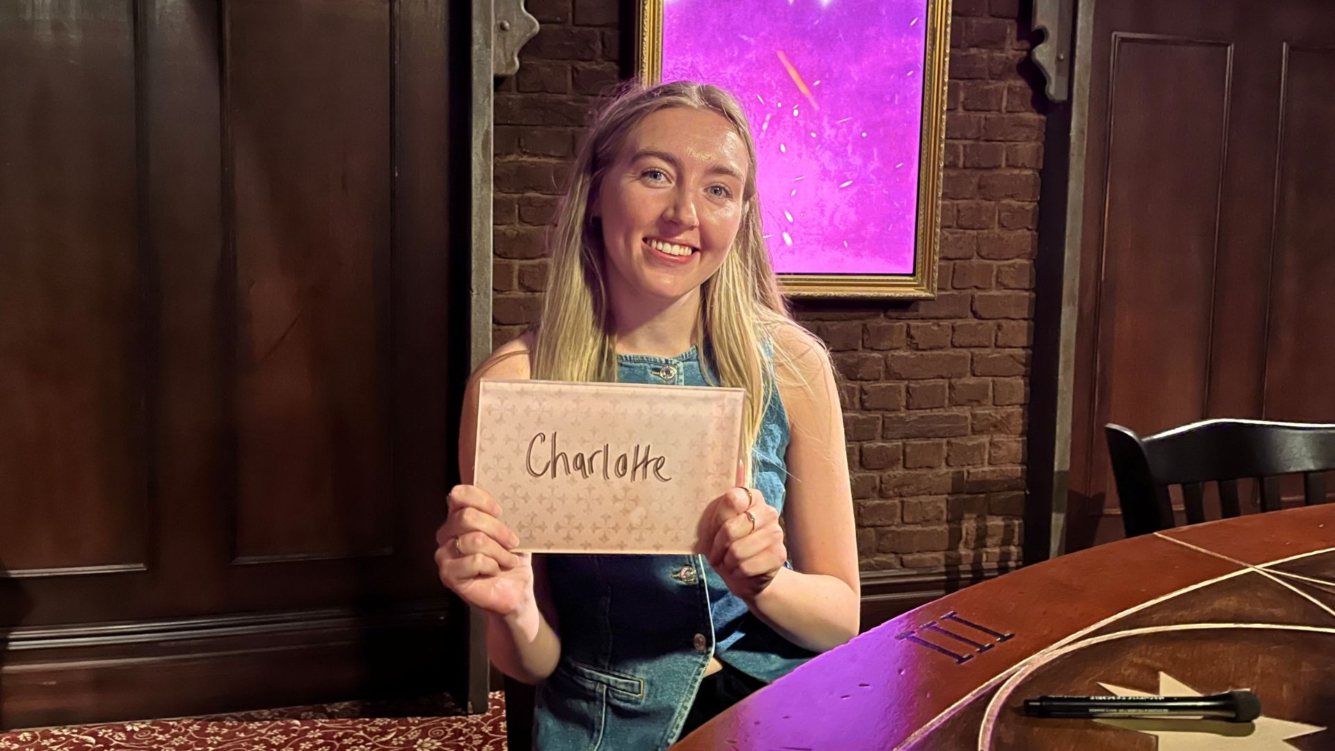 Proclaim your innocence as a Faithful at The Traitors: Live Experience. © Visit London/Charlotte Pritchard. Person sitting a table holding up a name card with interactive screen in background.