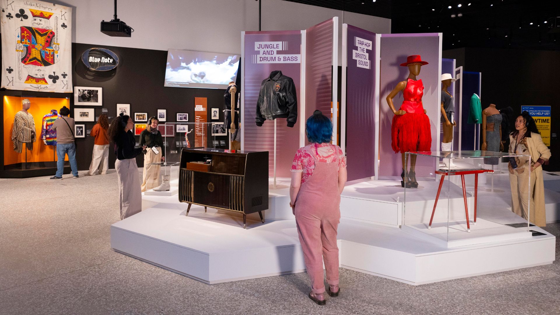 Explore V&A East Museum’s inaugural exhibition, The Music is Black: A British Story © David Parry for the V&A. Image courtesy of V&A East. Costumes and objects in centre of exhibition room with visitors looking at displays at The Music is Black exhibition.