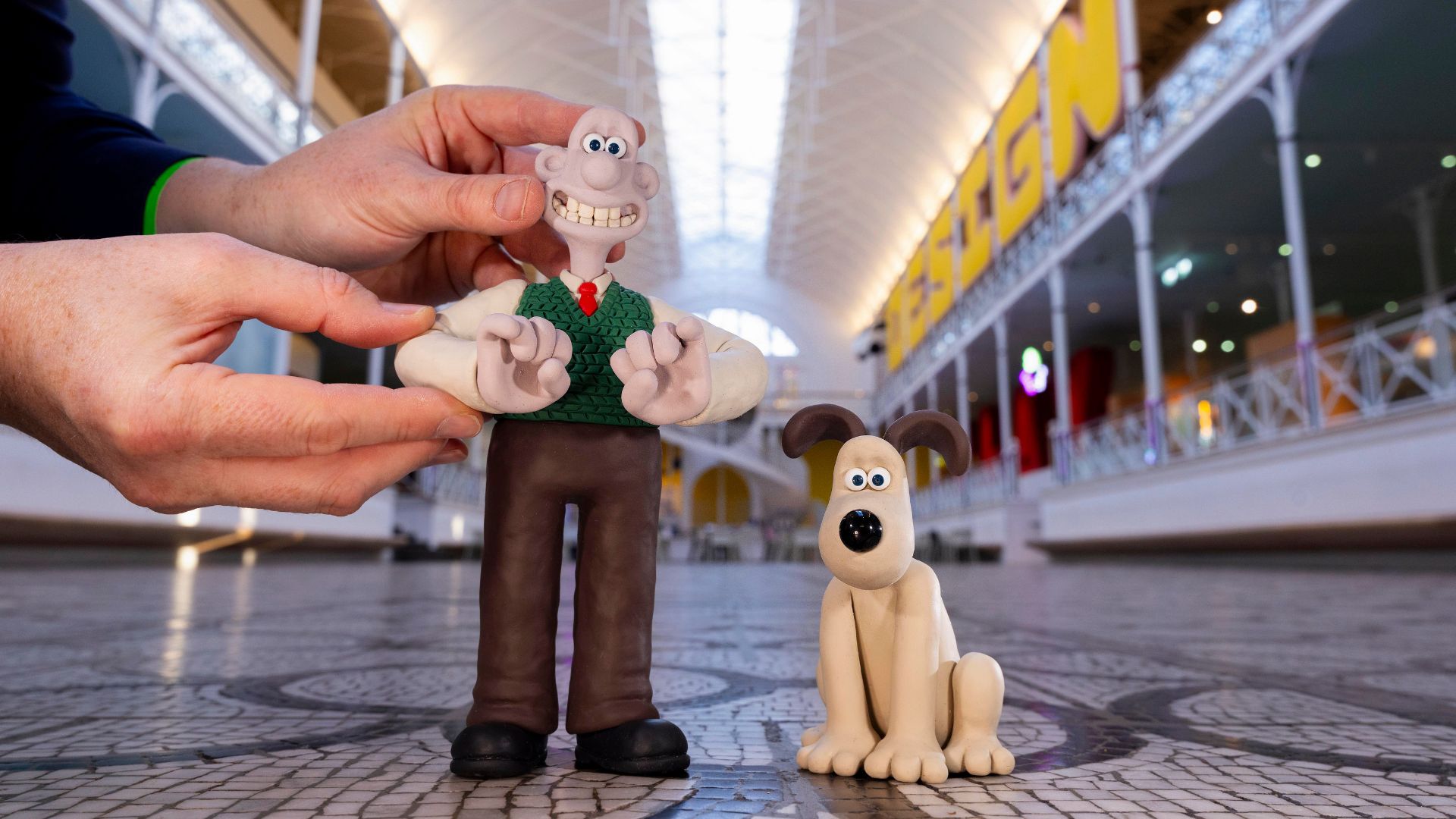 Behind the scenes of Wallace & Gromit’s photoshoot at Young V&A. Photo by David Parry for the V&A. Close-up of Wallace and Gromit clay figures being positioned on a patterned floor inside a bright, spacious gallery with high ceilings and a large “DESIGN” sign in the background.