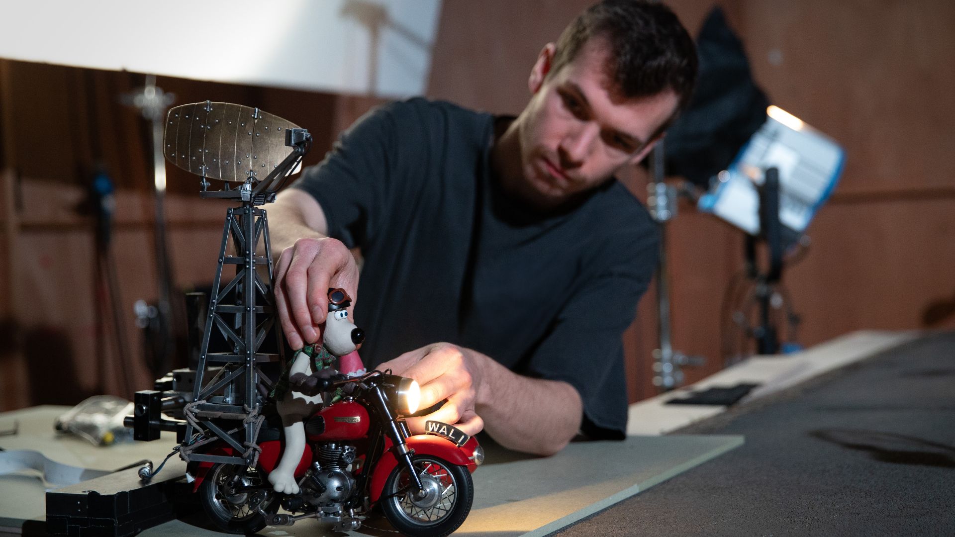 Production still from Wallace & Gromit: Vengeance Most Fowl (2024). Photo by Richard Davies. © TM Aardman Animations LTD/Wallace & Gromit LTD A person adjusts a detailed Wallace and Gromit motorcycle model on a workbench, surrounded by studio lighting and equipment.