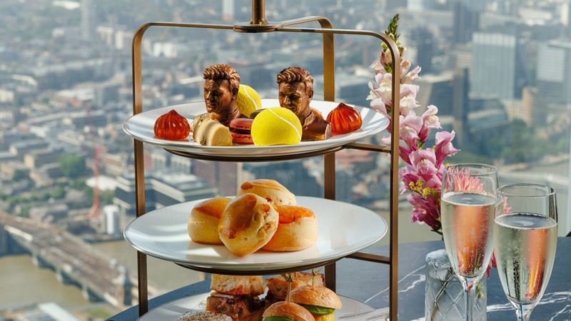 A Wimbledon-inspired afternoon tea served above the London skyline, where championship spirit meets breathtaking city views at Bread Street Kitchen & Bar by Gordon Ramsay. Image courtesy of Clementine Com. A Wimbledon-themed afternoon tea served on a three-tier stand beside floor-to-ceiling windows overlooking the London skyline. The spread includes tennis ball-shaped desserts, chocolate tennis player busts, freshly baked scones, savoury sandwiches, sparkling wine, and pink floral decorations.