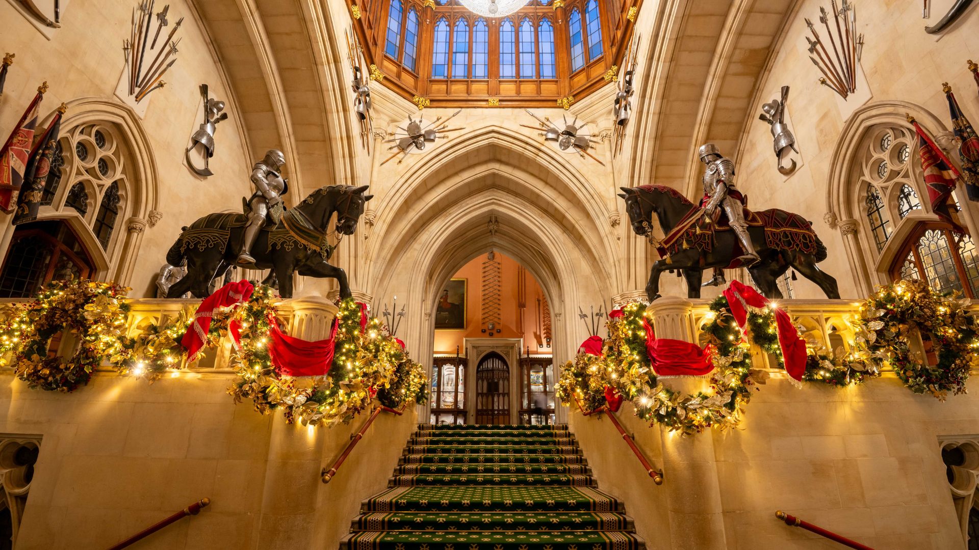 Ornate stone staircase at Windsor Castle decorated with festive garlands and red ribbons, flanked by two armored knight statues on horseback, with a vaulted ceiling and stained-glass windows above.