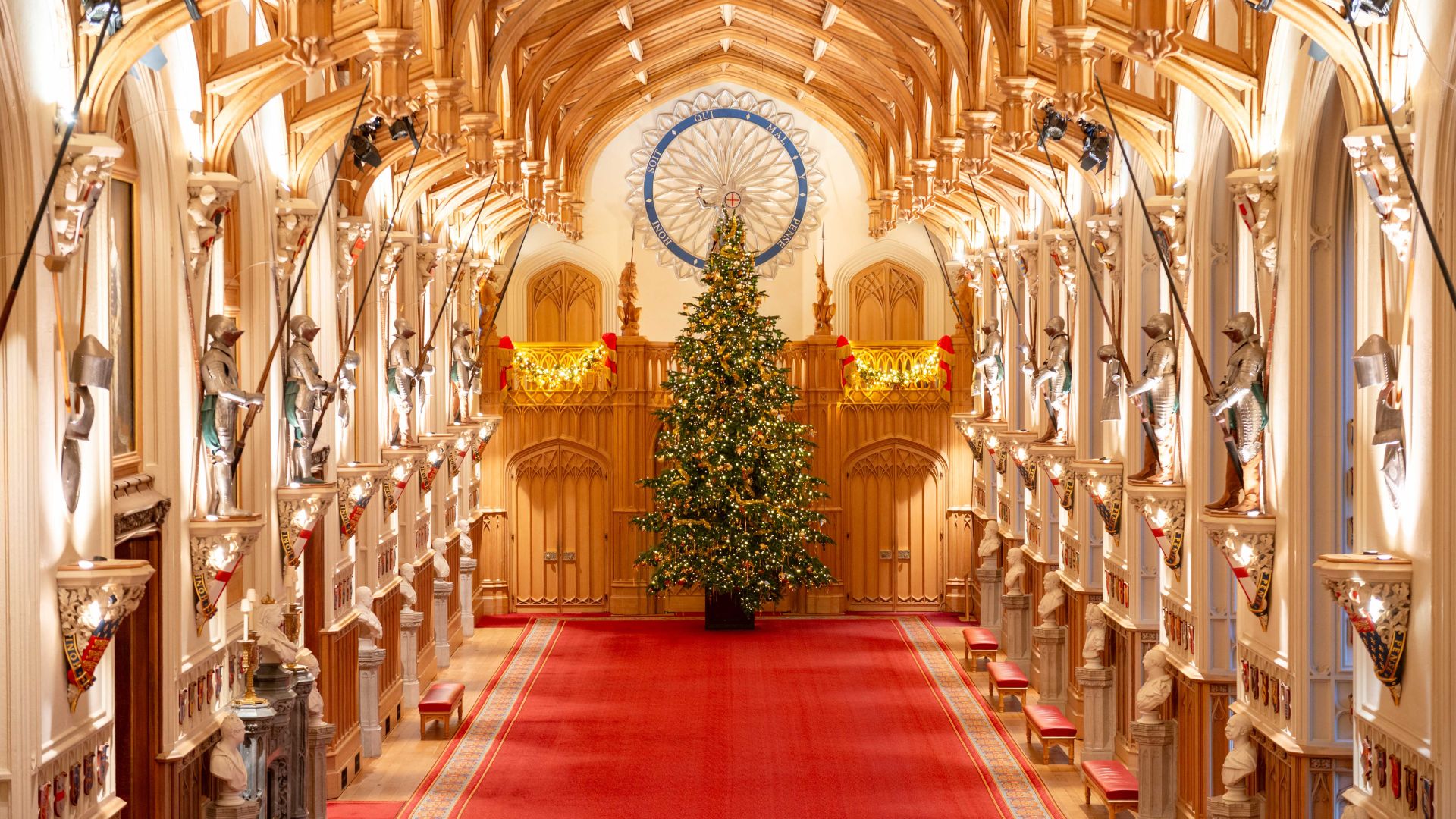 Find a 20ft Christmas tree in St George's Hall, Windsor Castle. © Royal Collection Enterprises Limited 2025 | Royal Collection Trust. St George’s Hall at Windsor Castle featuring a grand Christmas tree in the center, red carpet, and ornate wooden ceiling with suits of armour lining the walls.