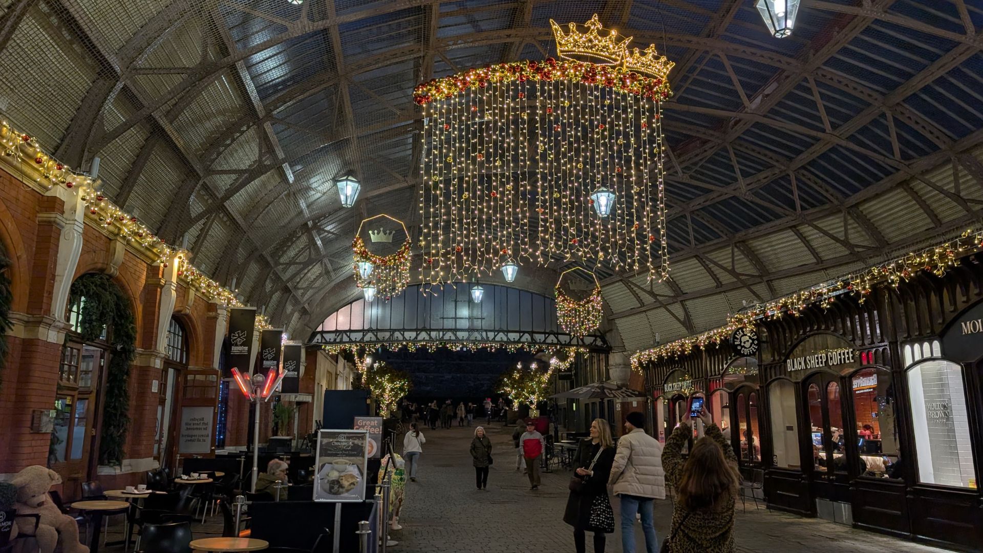 Covered shopping arcade adorned with Christmas lights and a large crown-shaped chandelier centerpiece, with people walking beneath the decorations.