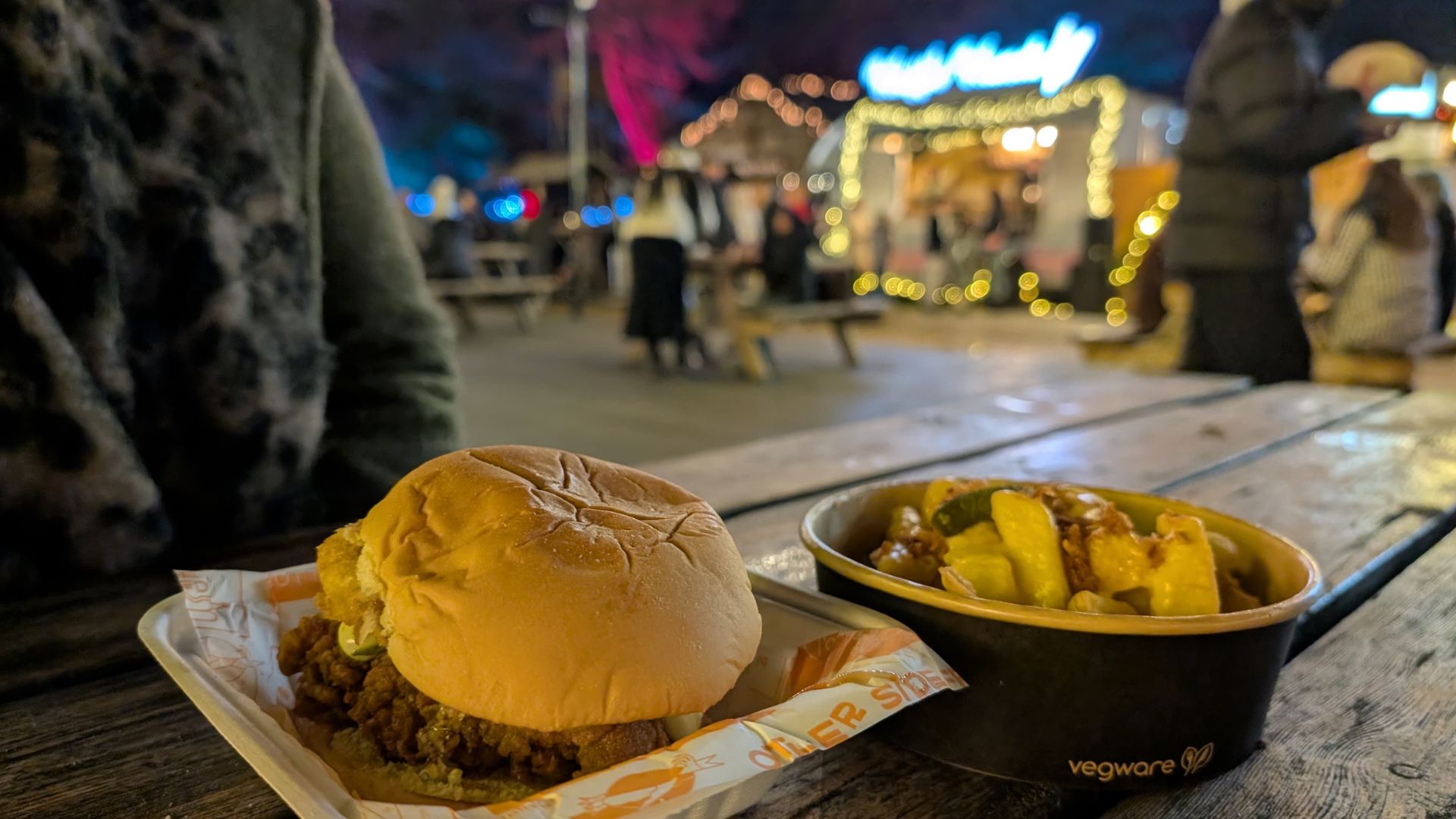 Close-up of a burger and a bowl of loaded fries on a wooden picnic table at a festive outdoor market, with blurred lights and food stalls in the background.