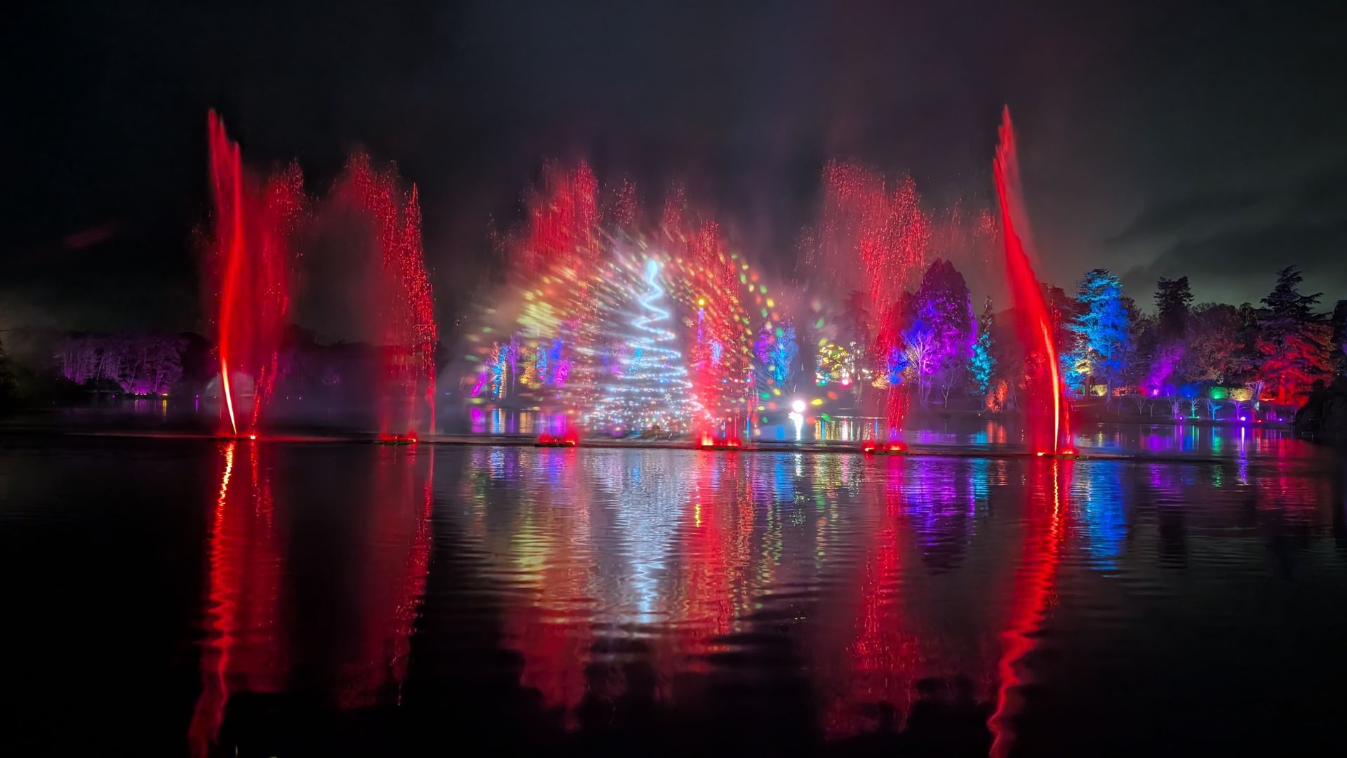 Experience spectacular light and water shows at Windsor Great Park Illuminated. Image courtesy of Hannah Cornish. Spectacular water fountain show with red jets and multicolored lights projected onto mist, reflecting on a calm lake at night.