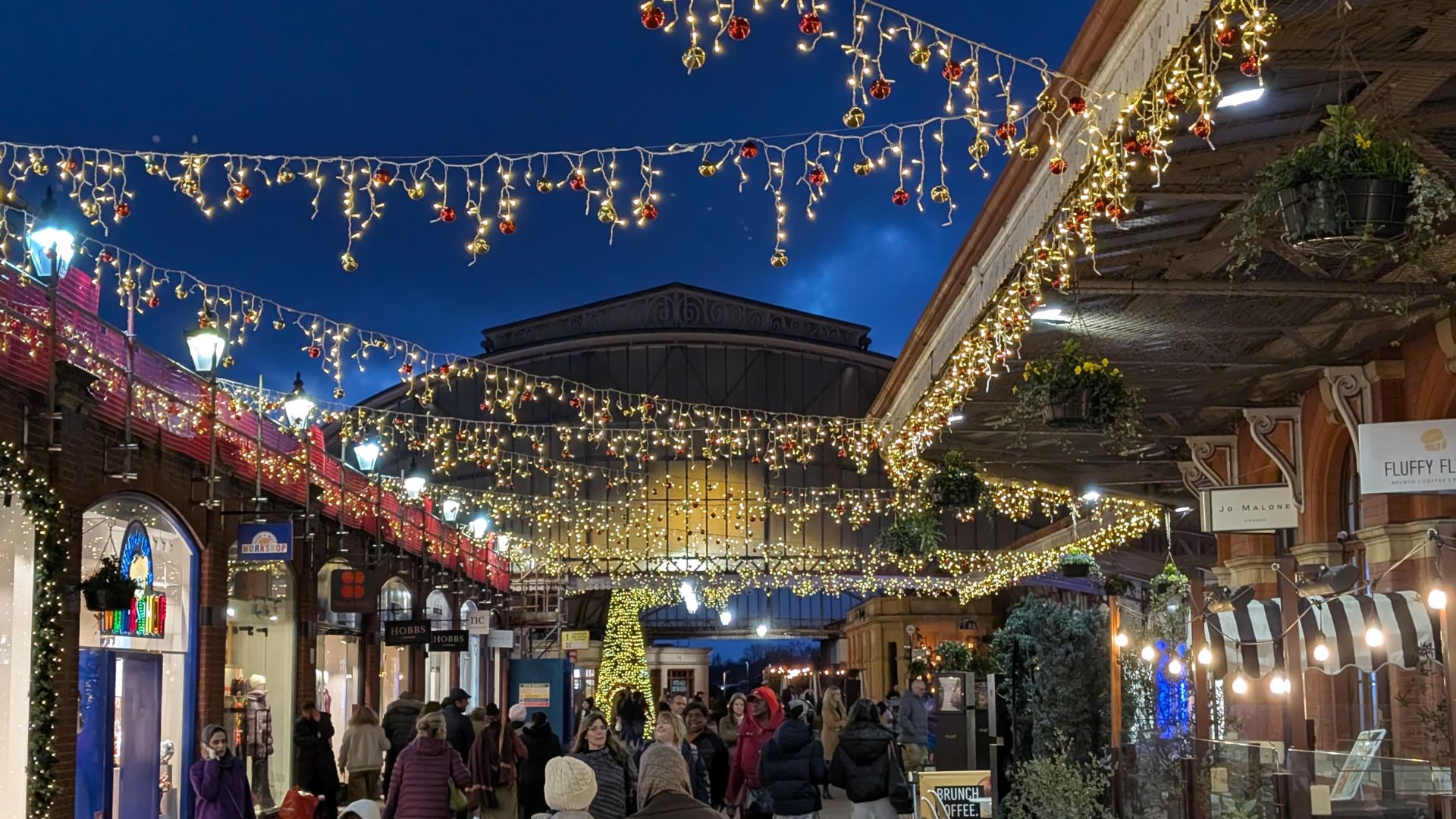 Outdoor shopping street decorated with strings of Christmas lights and ornaments, with crowds of people enjoying the festive atmosphere at dusk.