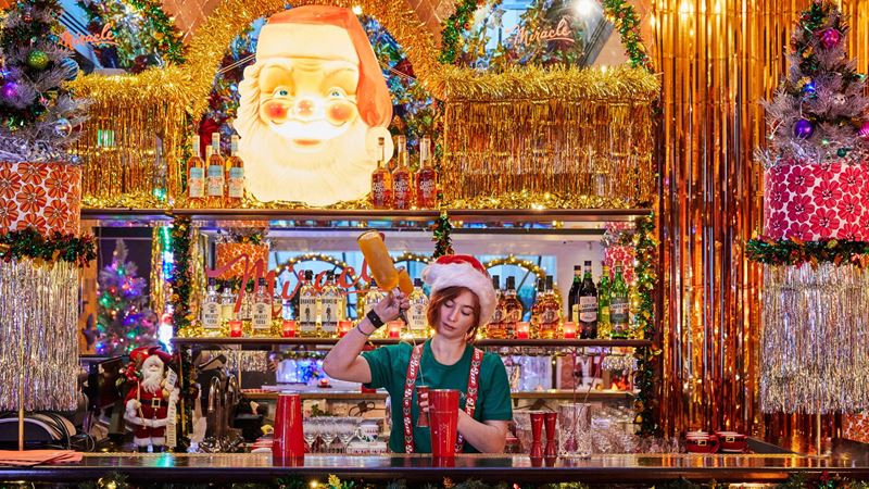 Decorated Christmas bar with tinsel and large Santa figure above the bar where a person dressed as an elf is pouring a drink.