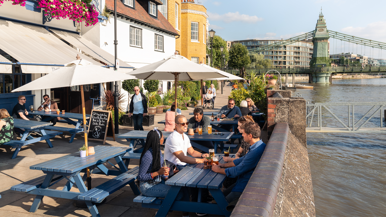 The Blue Anchor, Hammersmith. Image credit: Visit London/Antoine Buchet. Group of people enjoying drinks at a riverside pub in Hammersmith