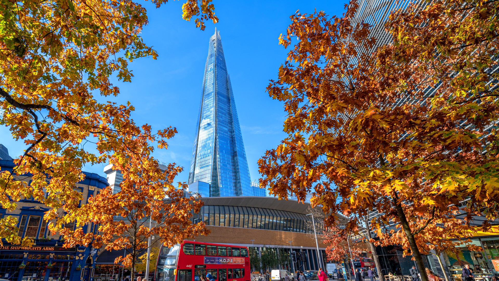 A photo of The Shard on a bright sunny day with a red bus and trees with autumn leaves in the foreground