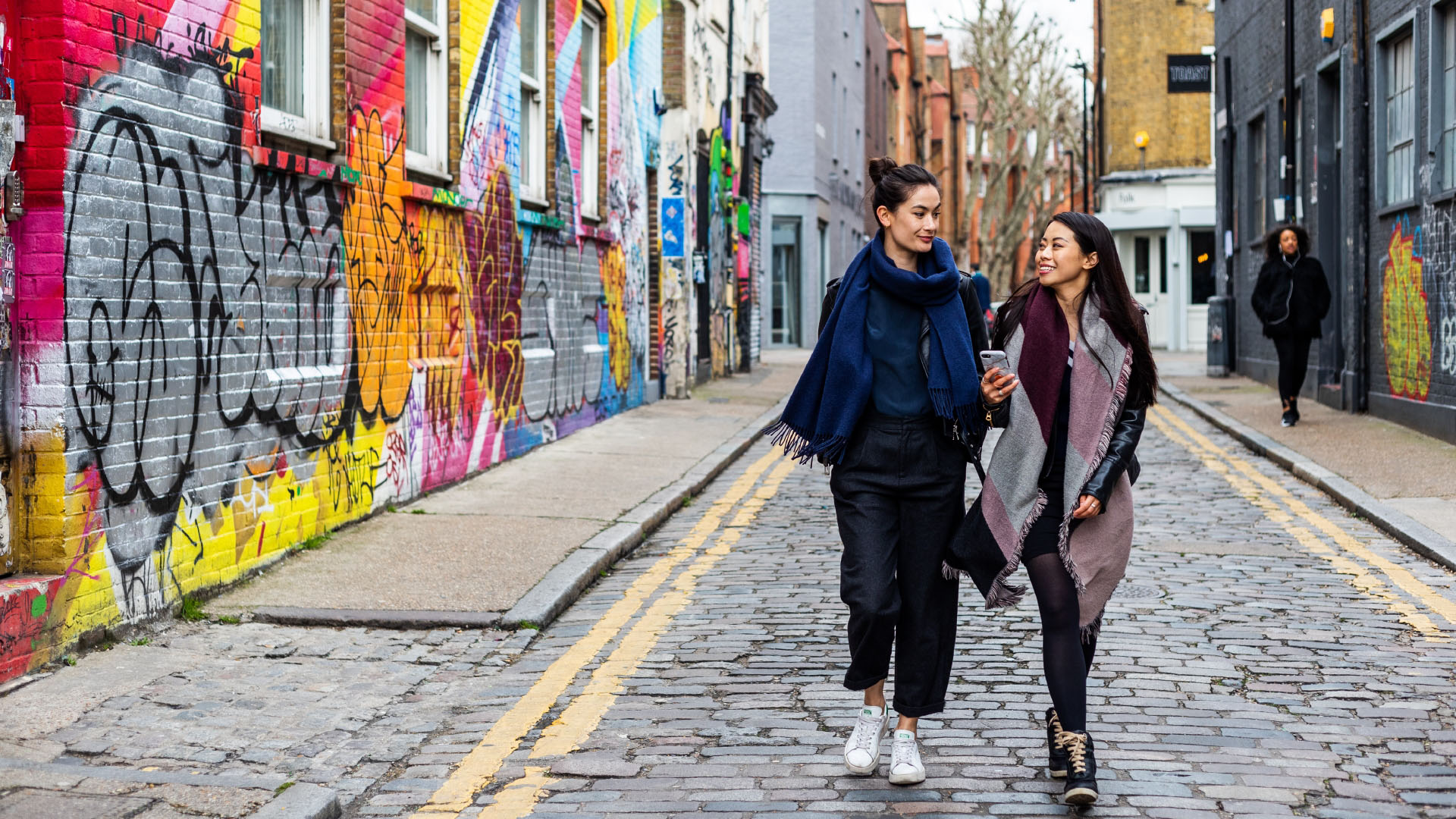 Découvrez les dernières œuvres de street art de l'est de Londres dans le quartier de Shoreditch. © London and Partners/Jon Reid. Deux femmes marchent dans une rue étroite en passant devant des œuvres de street art colorées à Shoreditch, dans l'est de Londres.