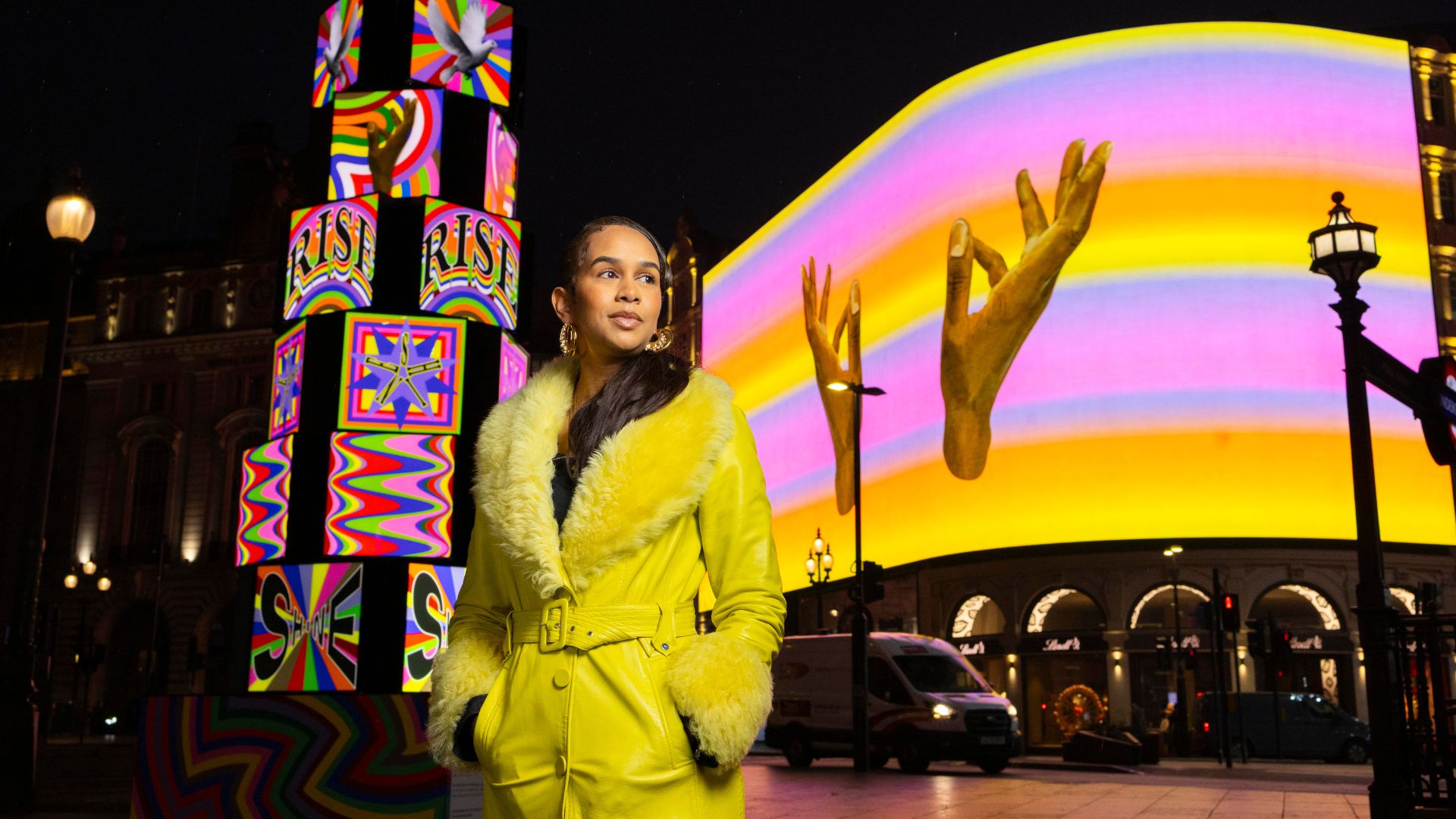 Artist Lakwena Maciver stands in front of her vibrant Art After Dark installation at Piccadilly Circus, where bold colour, movement and messages of joy transform London’s most iconic screens after sunset. Image courtesy of Prmeier Comms. Artist Lakwena Maciver wearing a bright yellow coat stands at Piccadilly Circus at night, surrounded by colourful digital art installations and illuminated billboards displaying rainbow patterns and symbolic imagery.